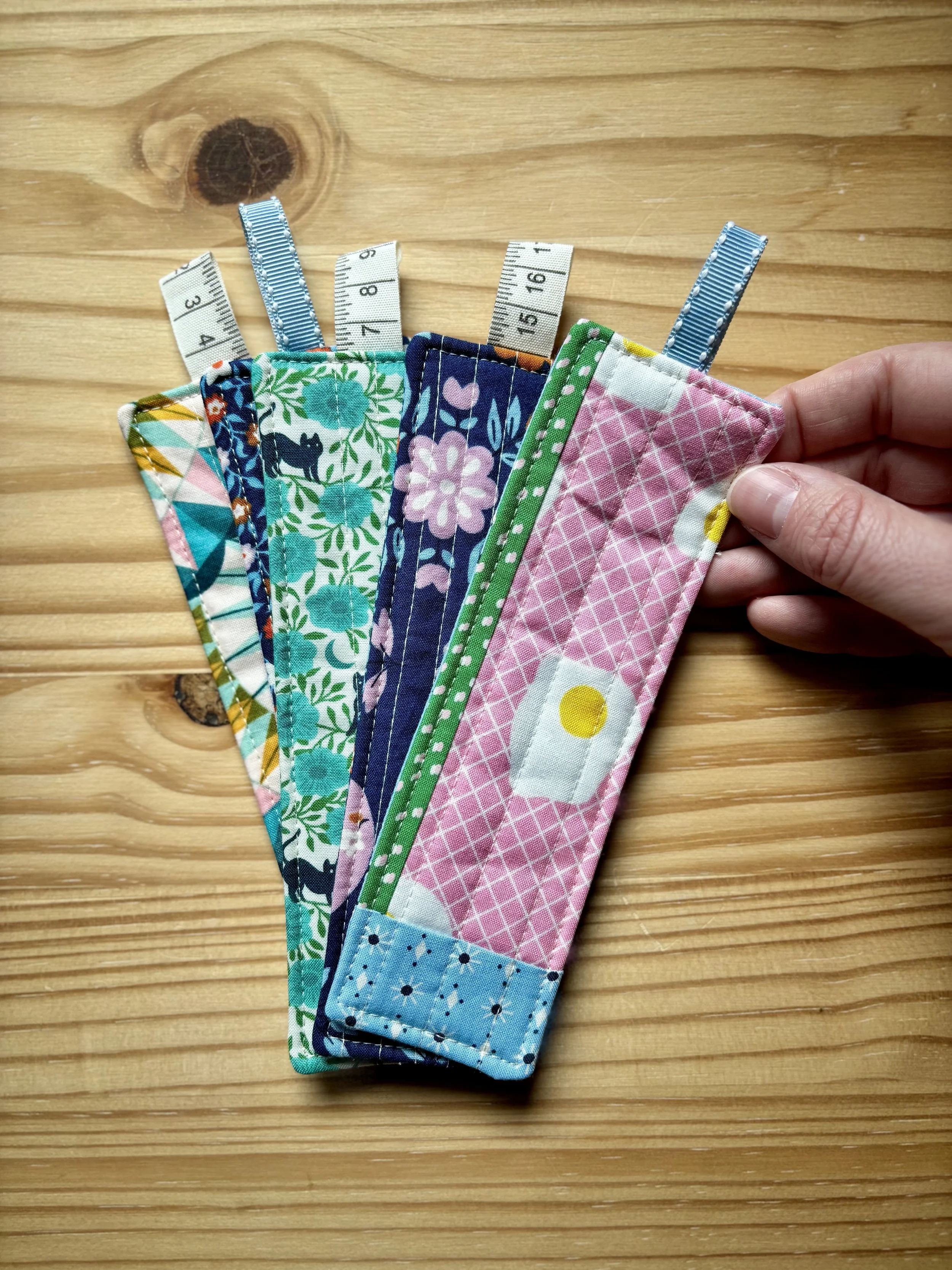 Image of 5 completed quilted bookmarks fanned out. Made using various Ruby Star Society prints. You can see a hand holding one of them as they rest on a natural pine wood surface.
