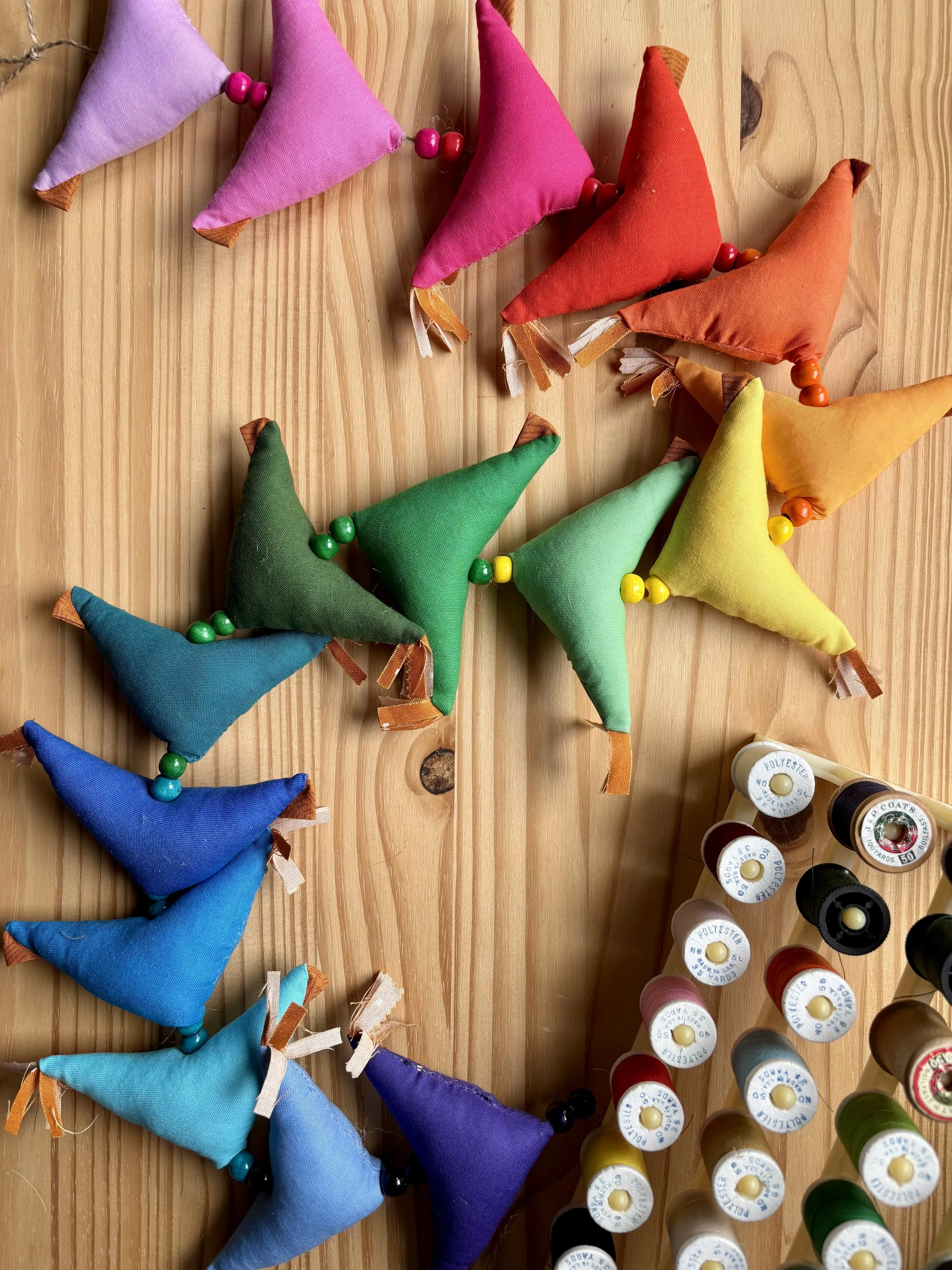 A bird's eye view of a rainbow colour gradient strand of prosperity hens in a curvy letter 'S' formation laying on a natural pine wood surface. A thread stand with various colour threads is also visible.