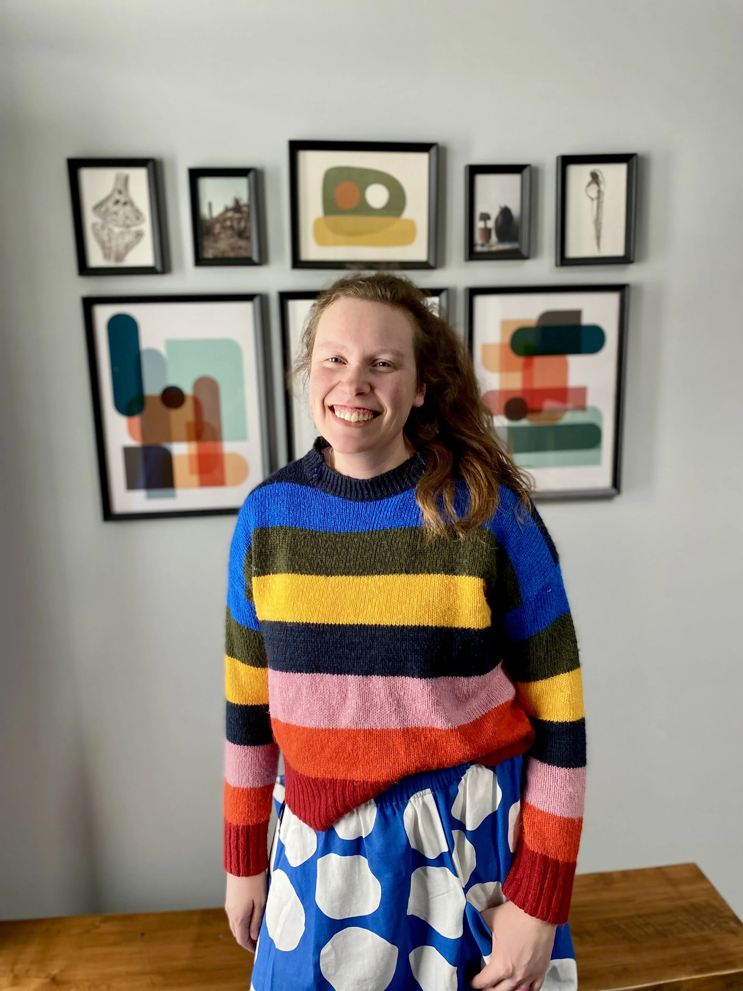 A photo of Natasha wearing a striped rainbow knit sweater smiling in front of a gallery wall of photos in a room with natural light.