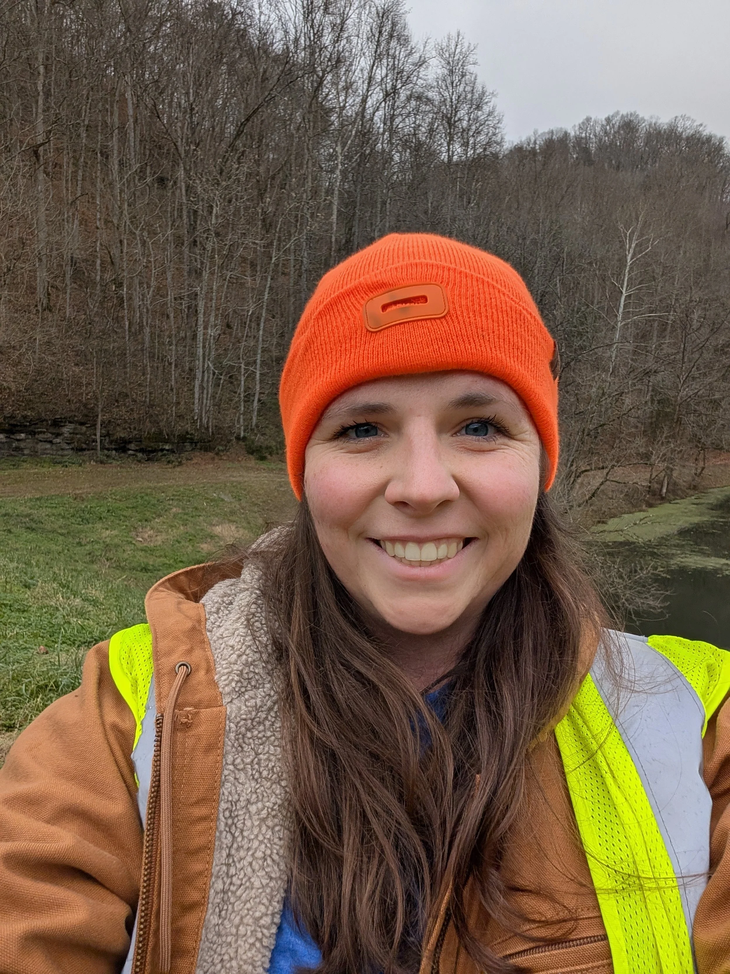 A woman outdoors wearing an orange beanie, a tan jacket with a yellow safety vest, and a blue shirt, smiling with a forest and a body of water in the background.