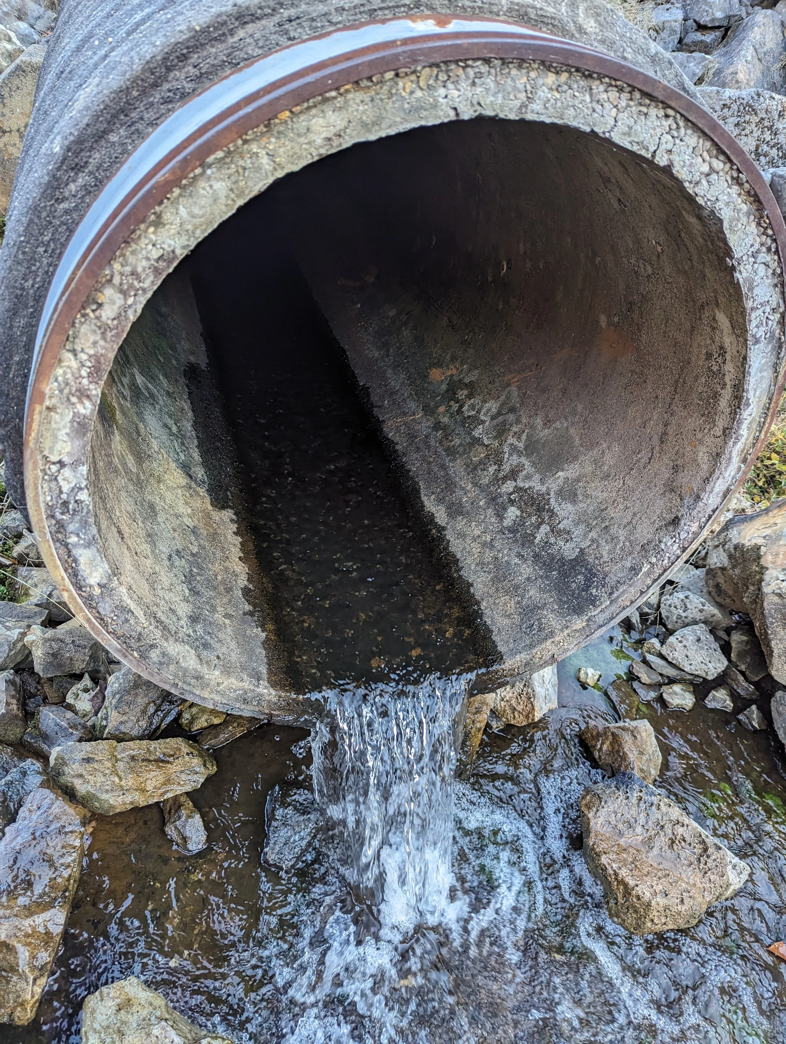 Water flowing out of a large, rusted metal pipe onto rocky ground.