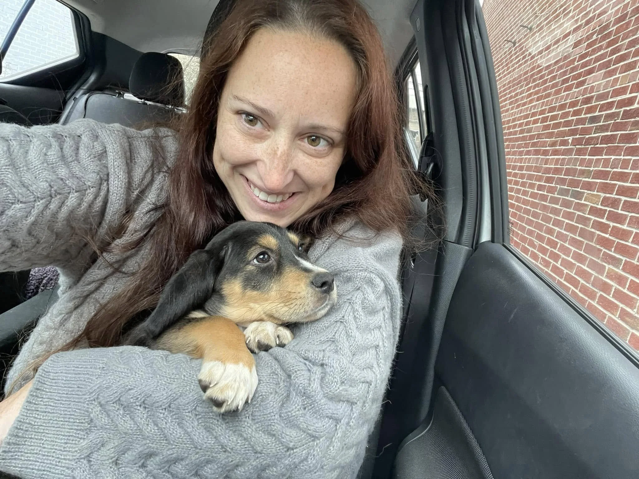A woman with reddish-brown hair and light skin smiling while holding a small black and tan puppy in her arms inside a vehicle with a brick wall outside the window.