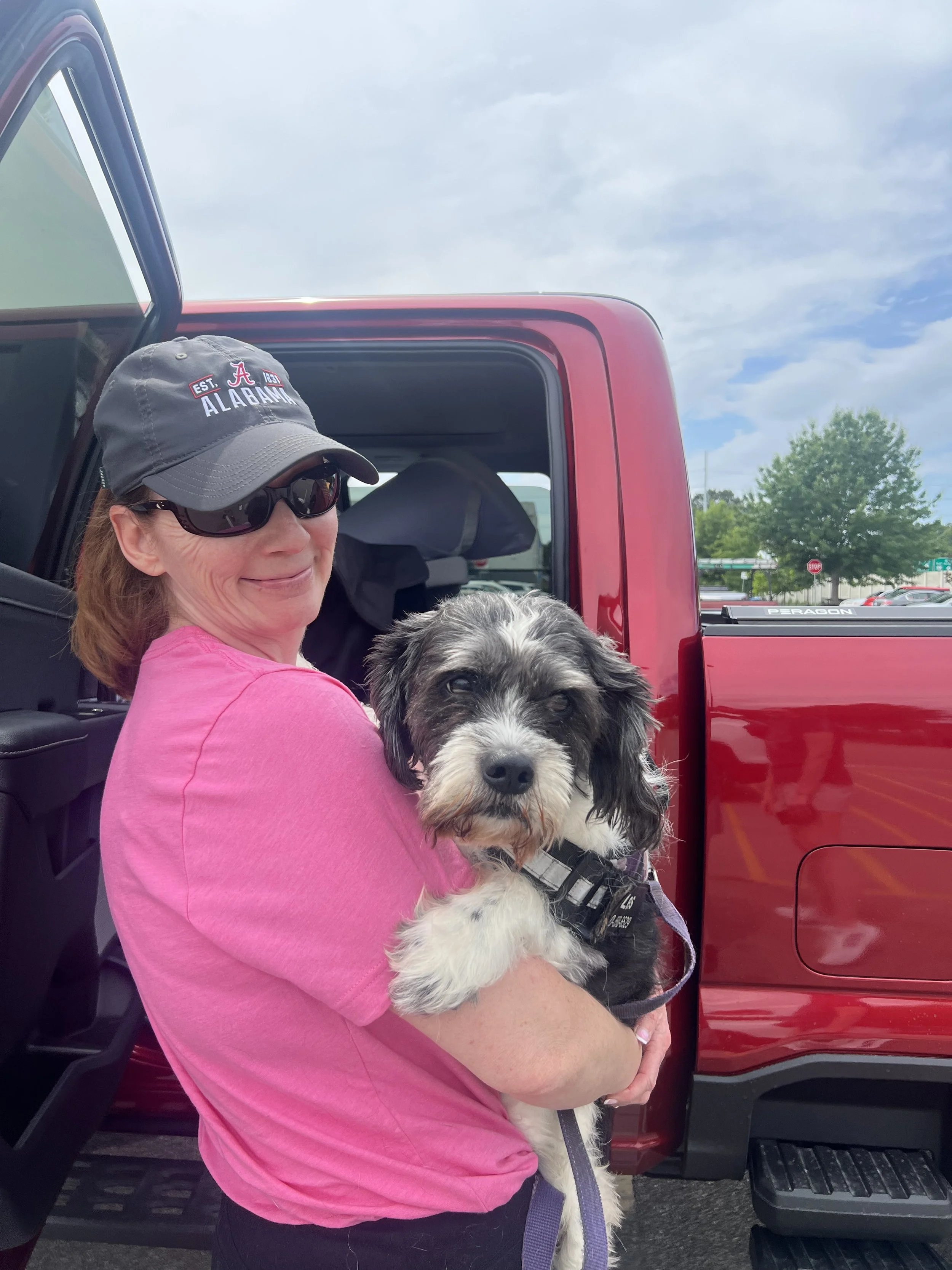 A woman holding a dog outside in front of a red pickup truck. The woman is wearing sunglasses, a gray cap with 'Alabama' written on it, and a pink shirt. The dog appears to be a black and white fluffy breed wearing a harness.