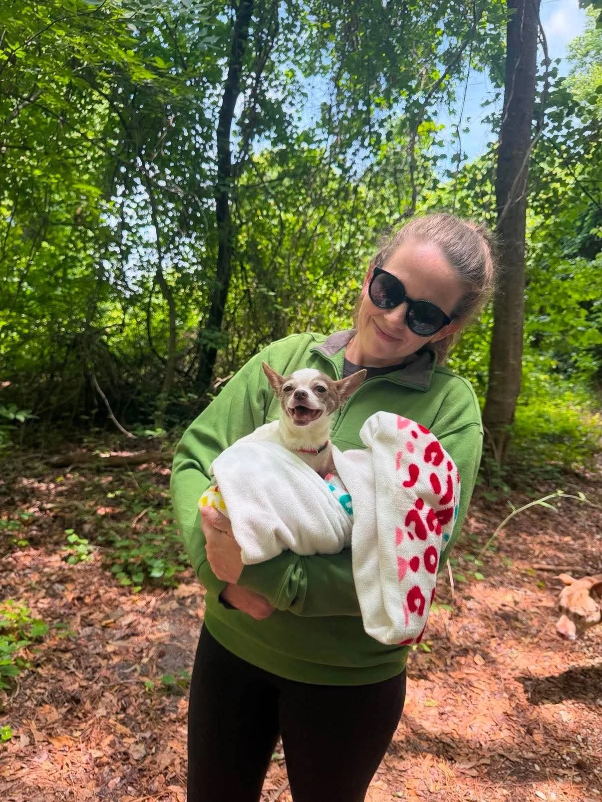 A woman wearing sunglasses and a green jacket is holding a small dog wrapped in a white towel with pink and red writing, in a wooded area with trees and foliage in the background. The dog looks happy and smiling.