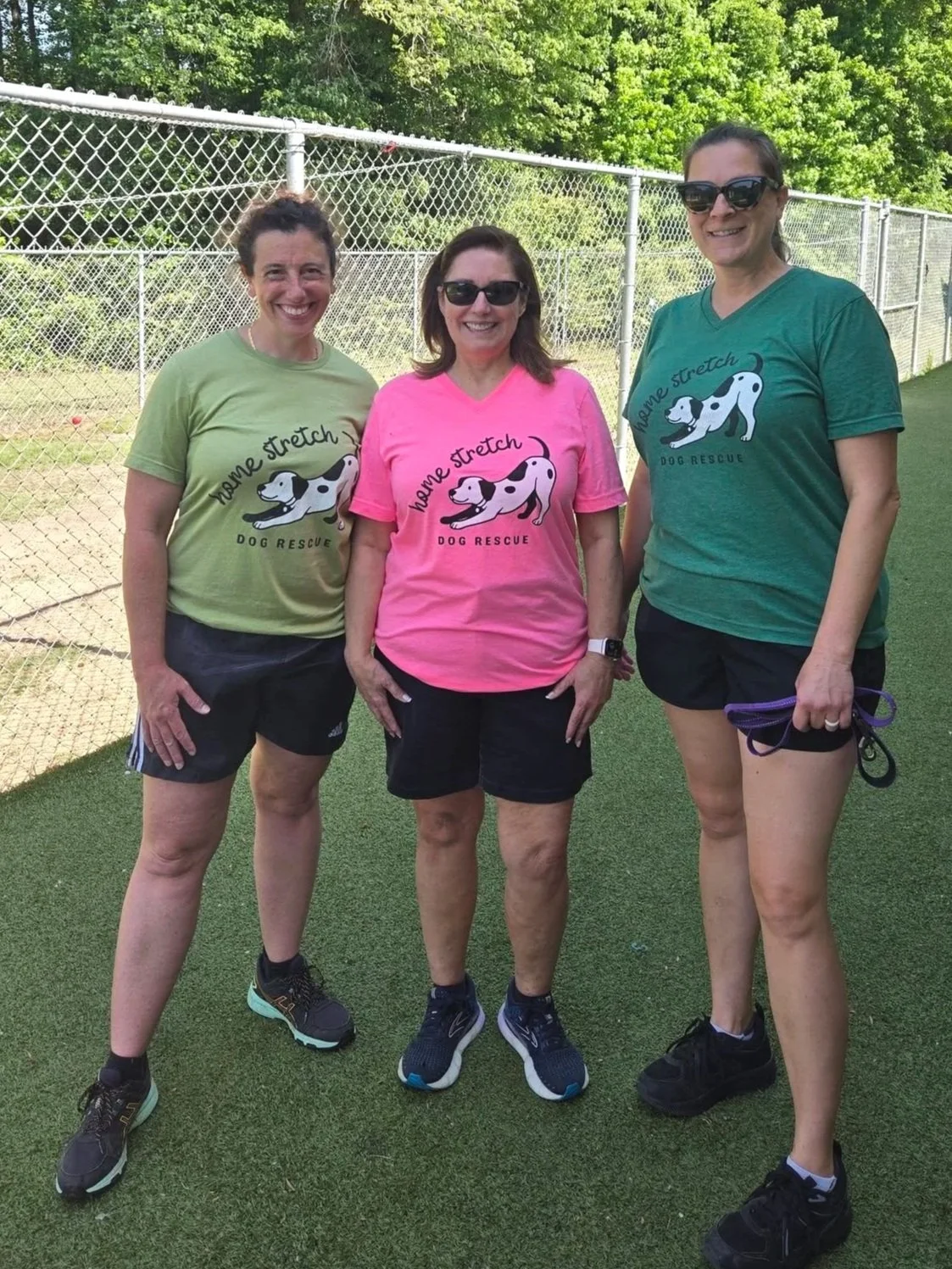 Three women standing outdoors on a grassy field near a chain-link fence, wearing colorful t-shirts with a dog rescue logo, shorts, and athletic shoes, smiling at the camera.
