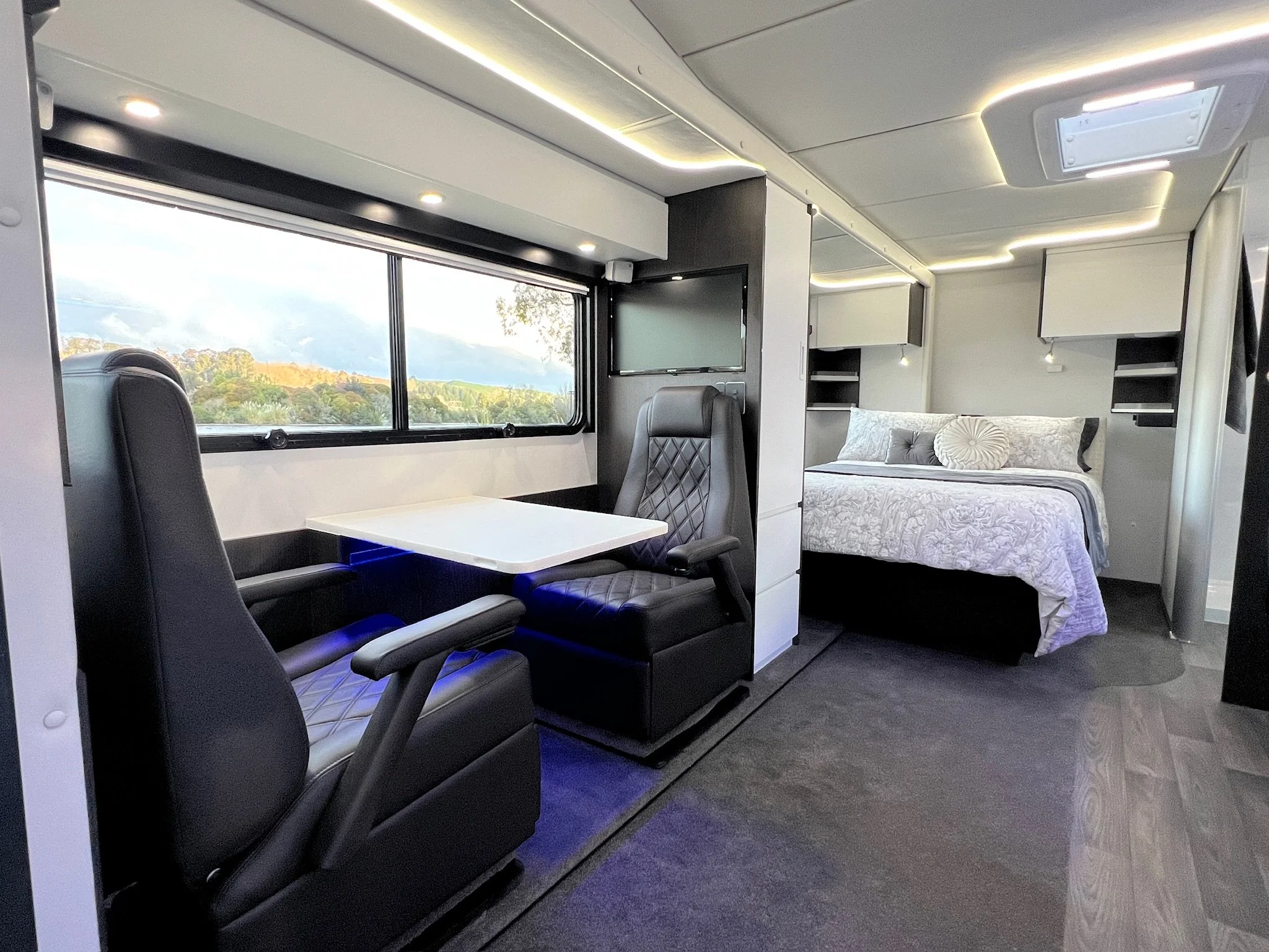Interior slide out of a modern mobile home or camper with a dining area, two black leather chairs, a white table, a large window showing a scenic outdoor view, a TV, and a bed with pillows and a white bedspread.