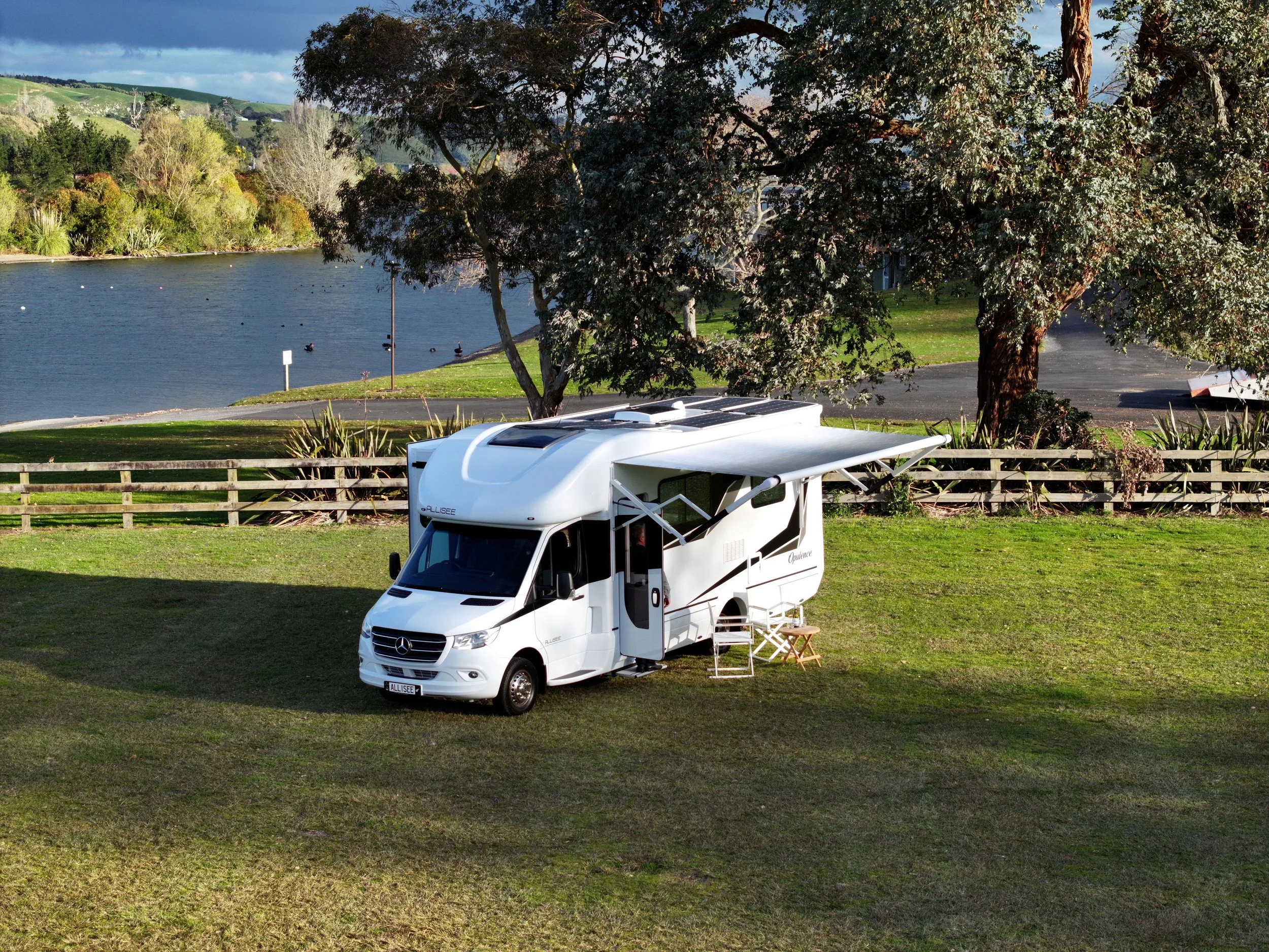 A white camper van parked on a grassy area near a lake with trees and hills in the background, along with outdoor chairs and a small table set up outside.