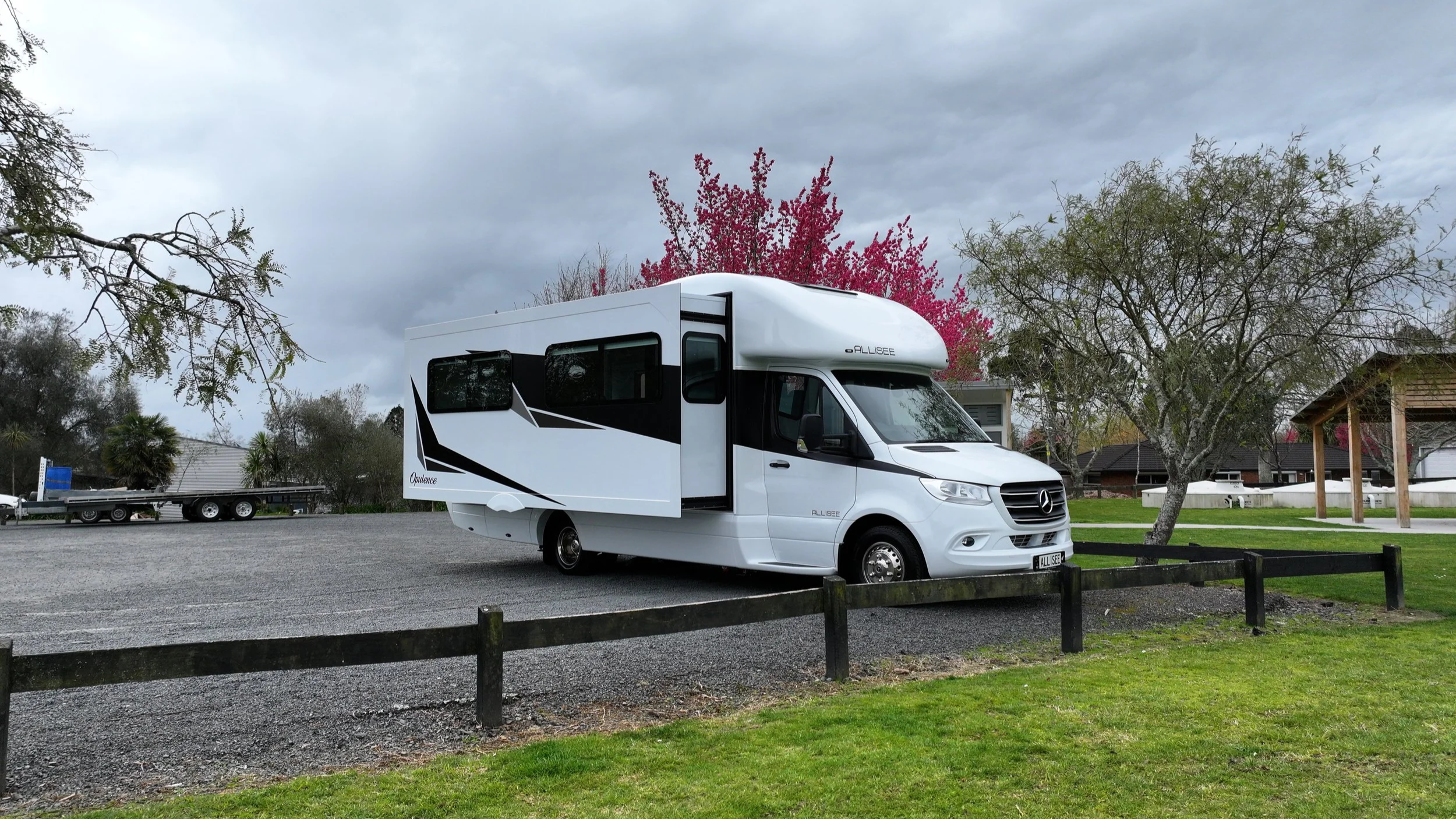 White motorhome parked in a grassy area with trees and residential houses in the background under a cloudy sky.