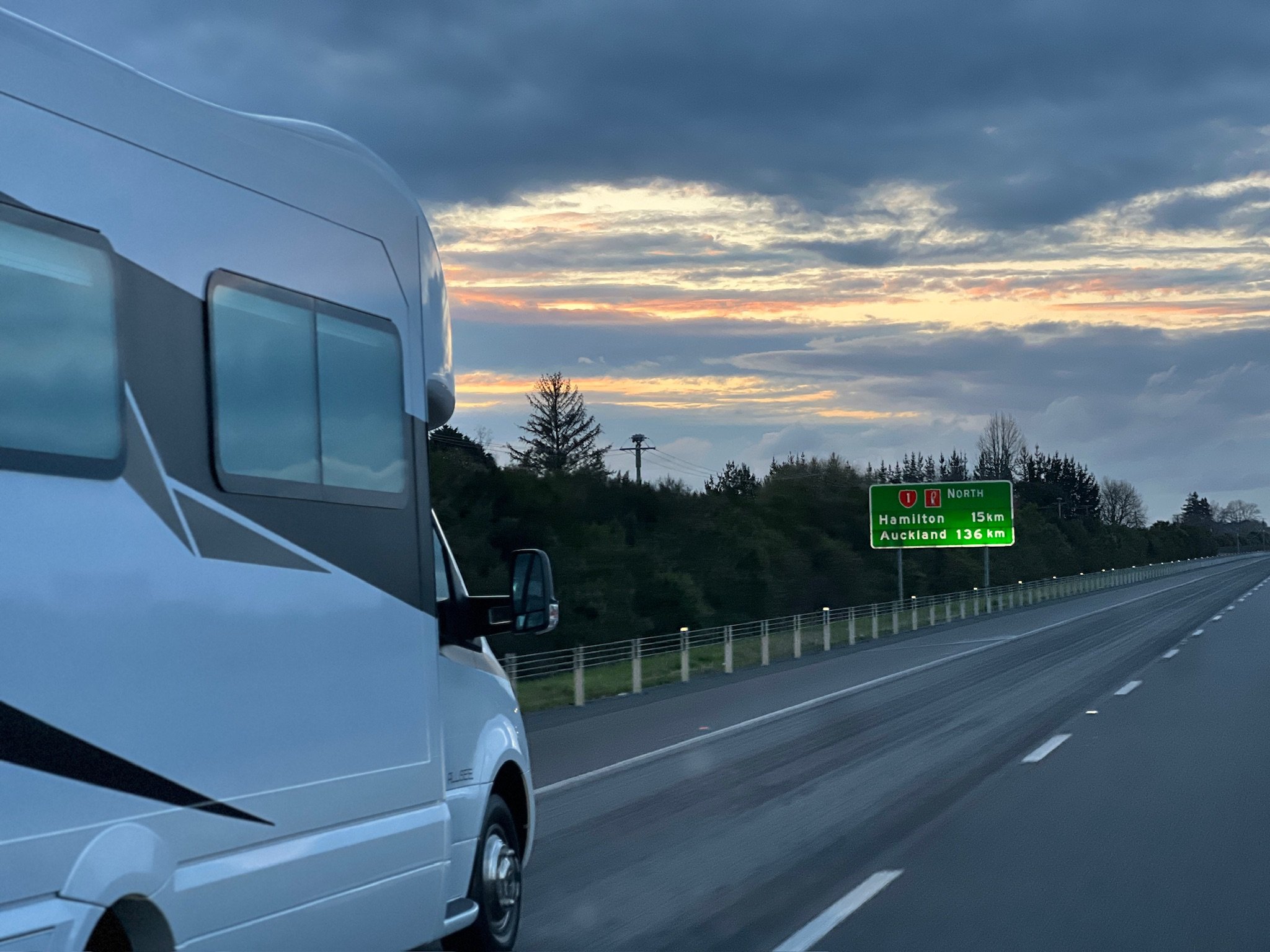 A white camper van on a highway during sunset. The sky is partly cloudy with streaks of orange and yellow. A green road sign indicates the distances to Hamilton (15 km) and Auckland (136 km).