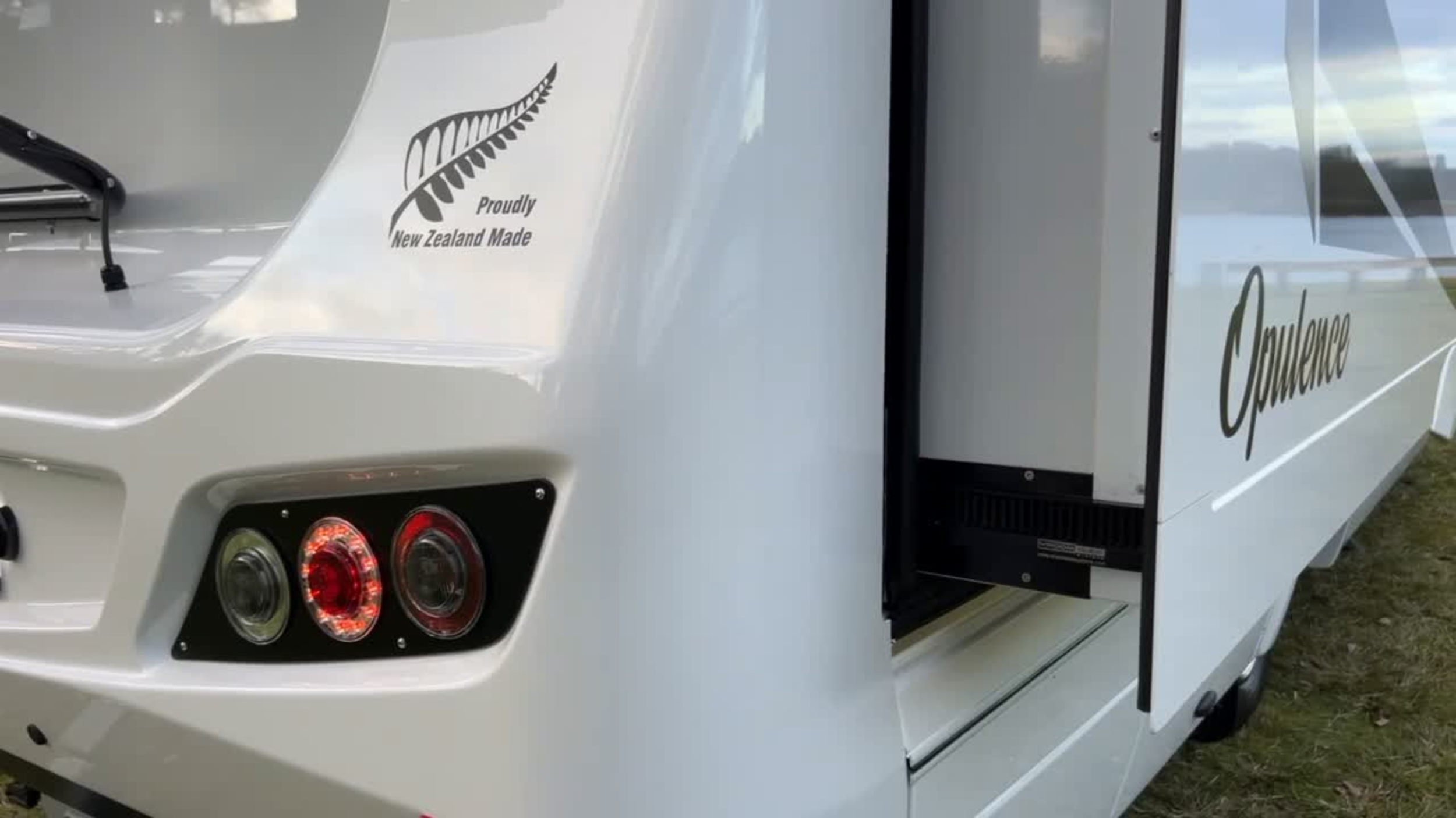 A white motorhome close up of the Vroom Slide Out system. The motorhome has a logo of a fern leaf and text that reads 'Proudly New Zealand Made' on the side and 'Opulence' on the door, parked on grass.
