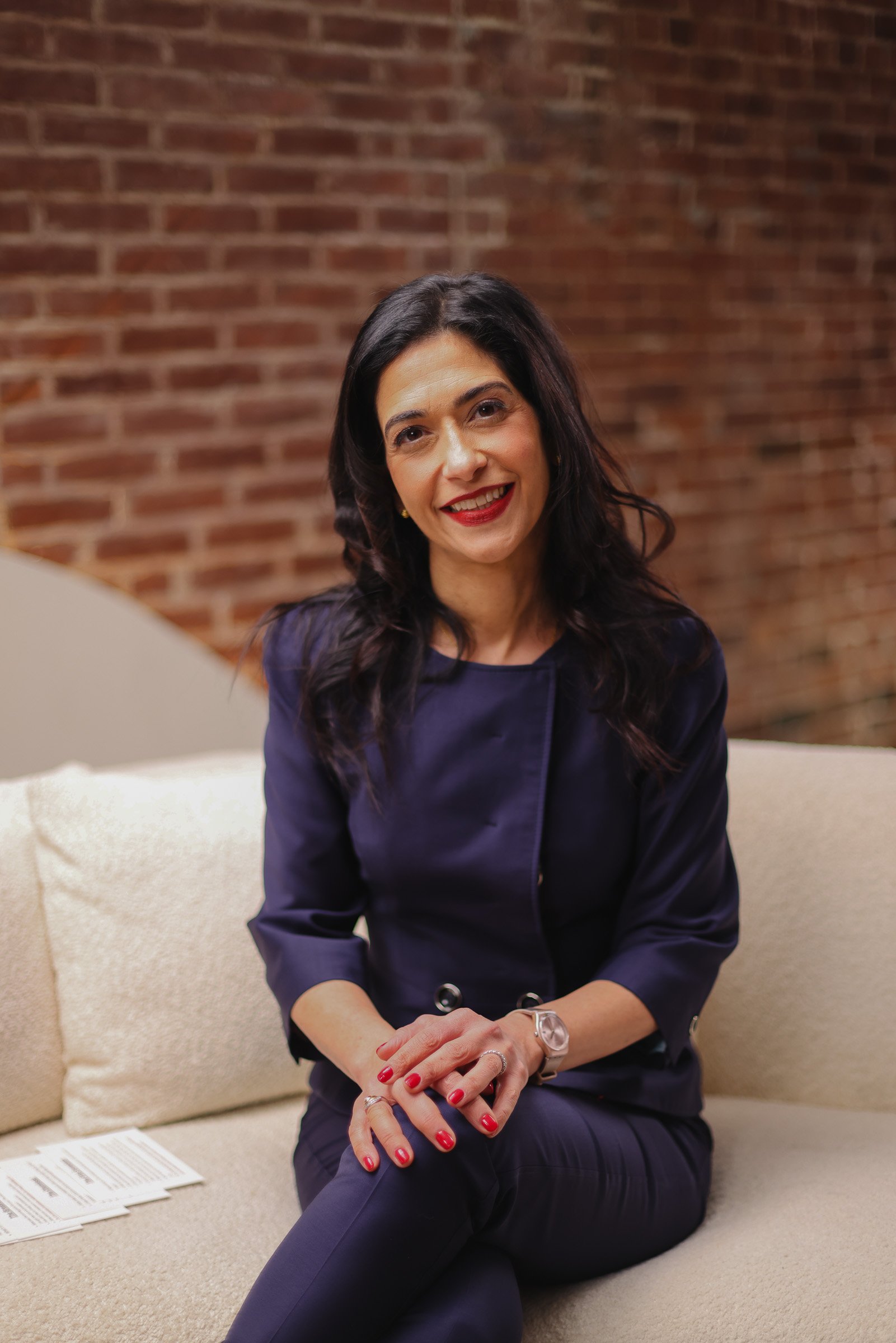 A woman with black hair, wearing a navy blue top and pants, sitting on a beige couch against a brick wall, smiling at the camera with red lipstick and red nail polish.