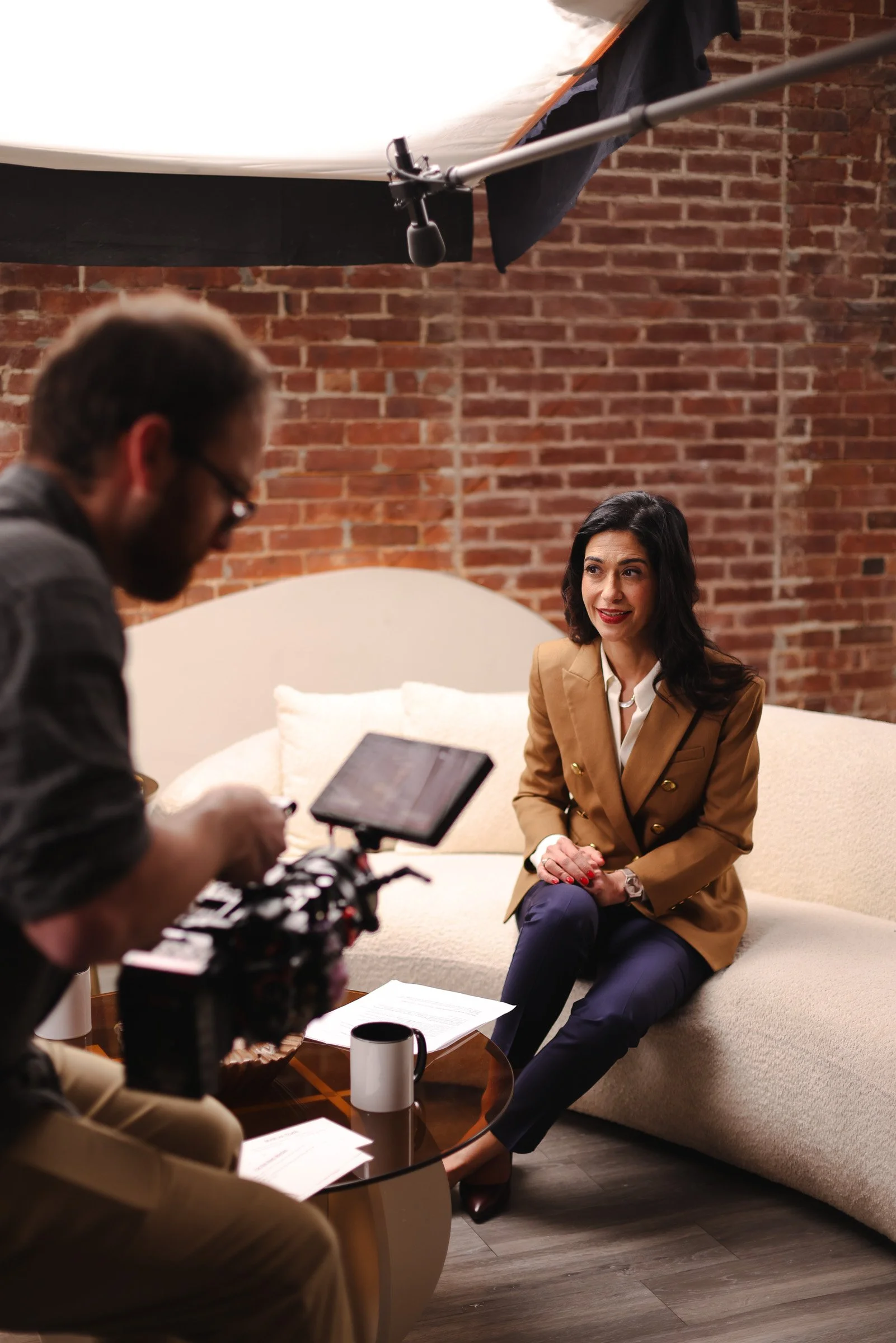 A woman is sitting on a cream curved sofa in an interview setting, wearing a tan blazer, white shirt, and navy pants, with a brick wall in the background. A man is filming her with professional equipment, holding a camera and standing beside a small glass table with papers, a mug, and a recording device, in a room with wooden flooring.