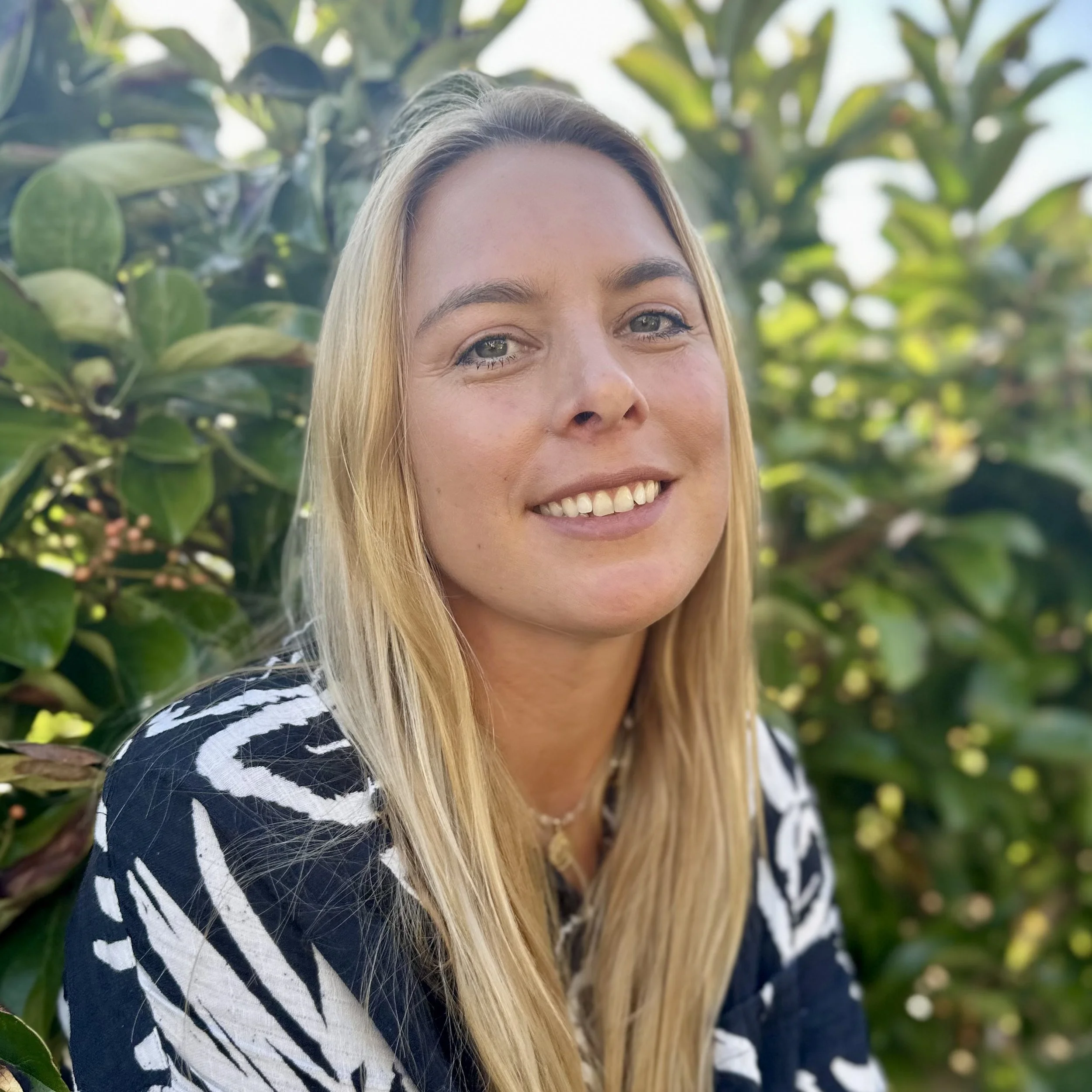 A smiling woman with long blonde hair posing outdoors in front of green leafy bushes, wearing a black and white patterned top.