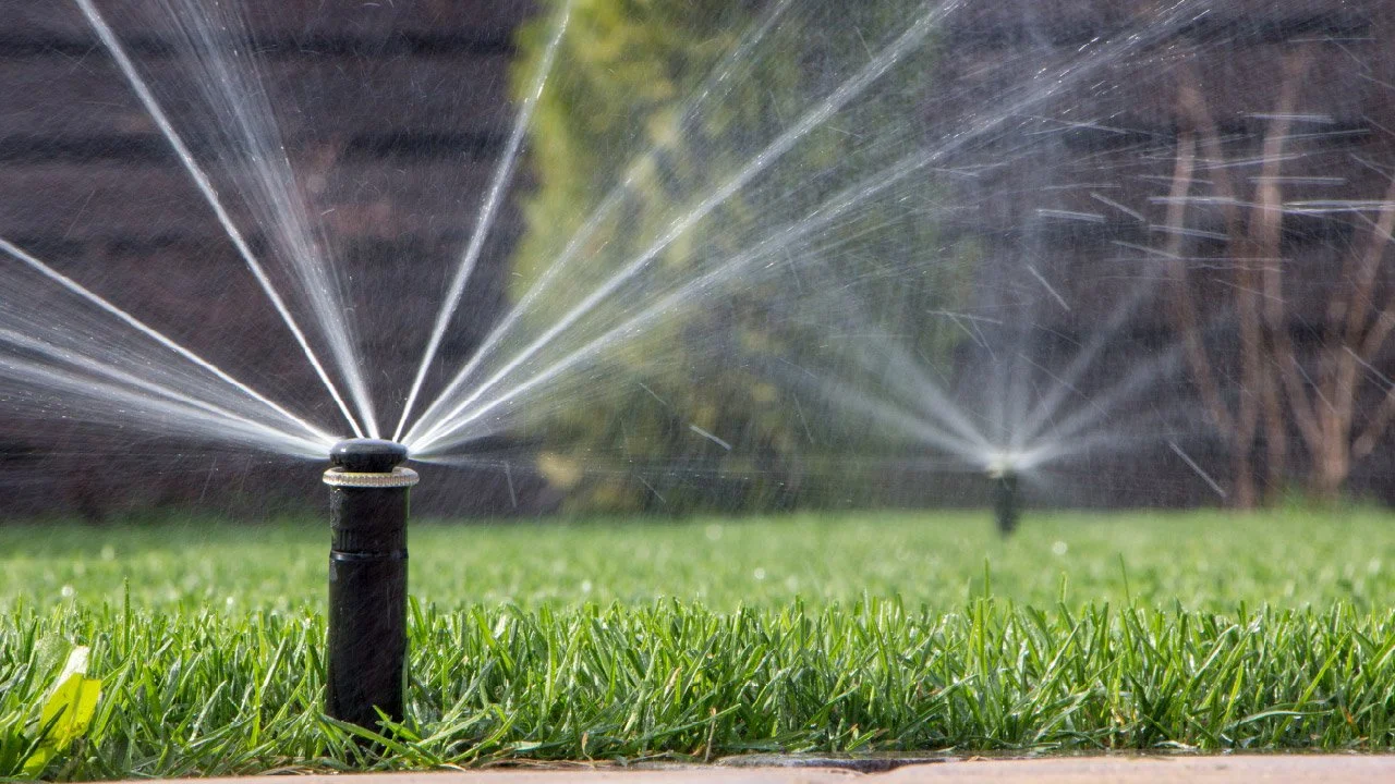 Close-up of a black sprinkler head watering green grass, with another sprinkler in the background watering more grass in a garden or yard.