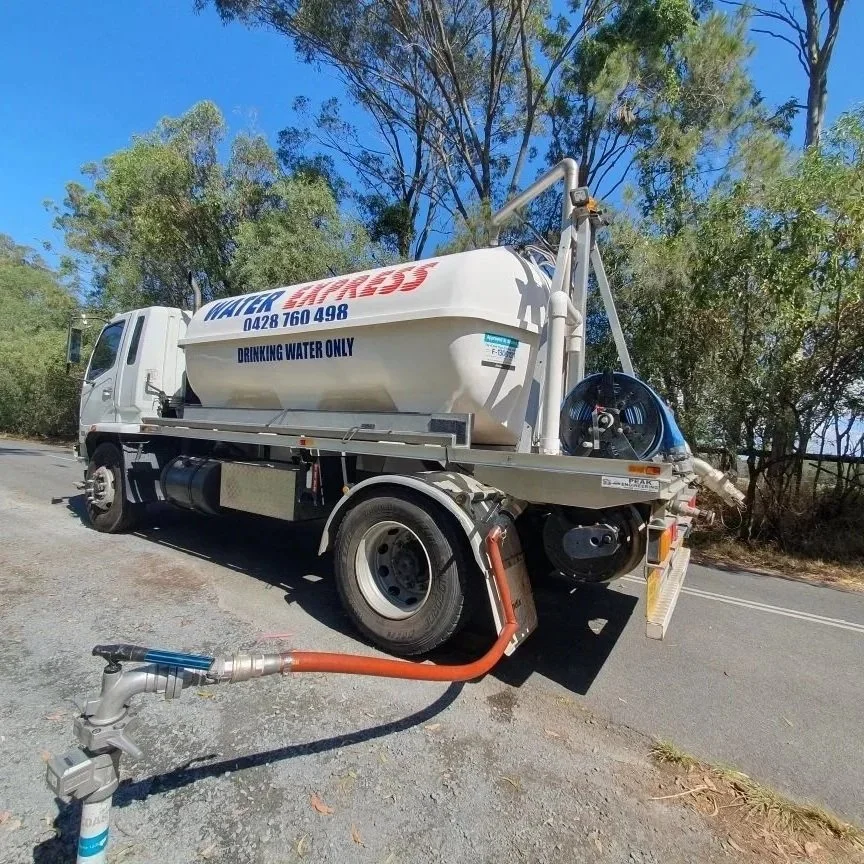 Water truck with a large tank labeled for drinking water, parked on a rural road. A hose extends from the truck to the ground.