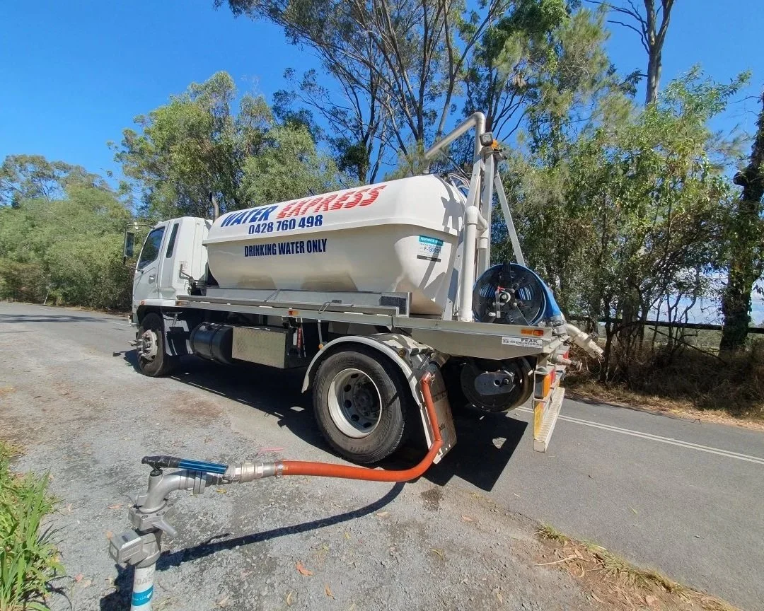 A water tanker truck parked on the side of a road, with a hose connected to a hydrant, in a rural area with trees and blue sky.