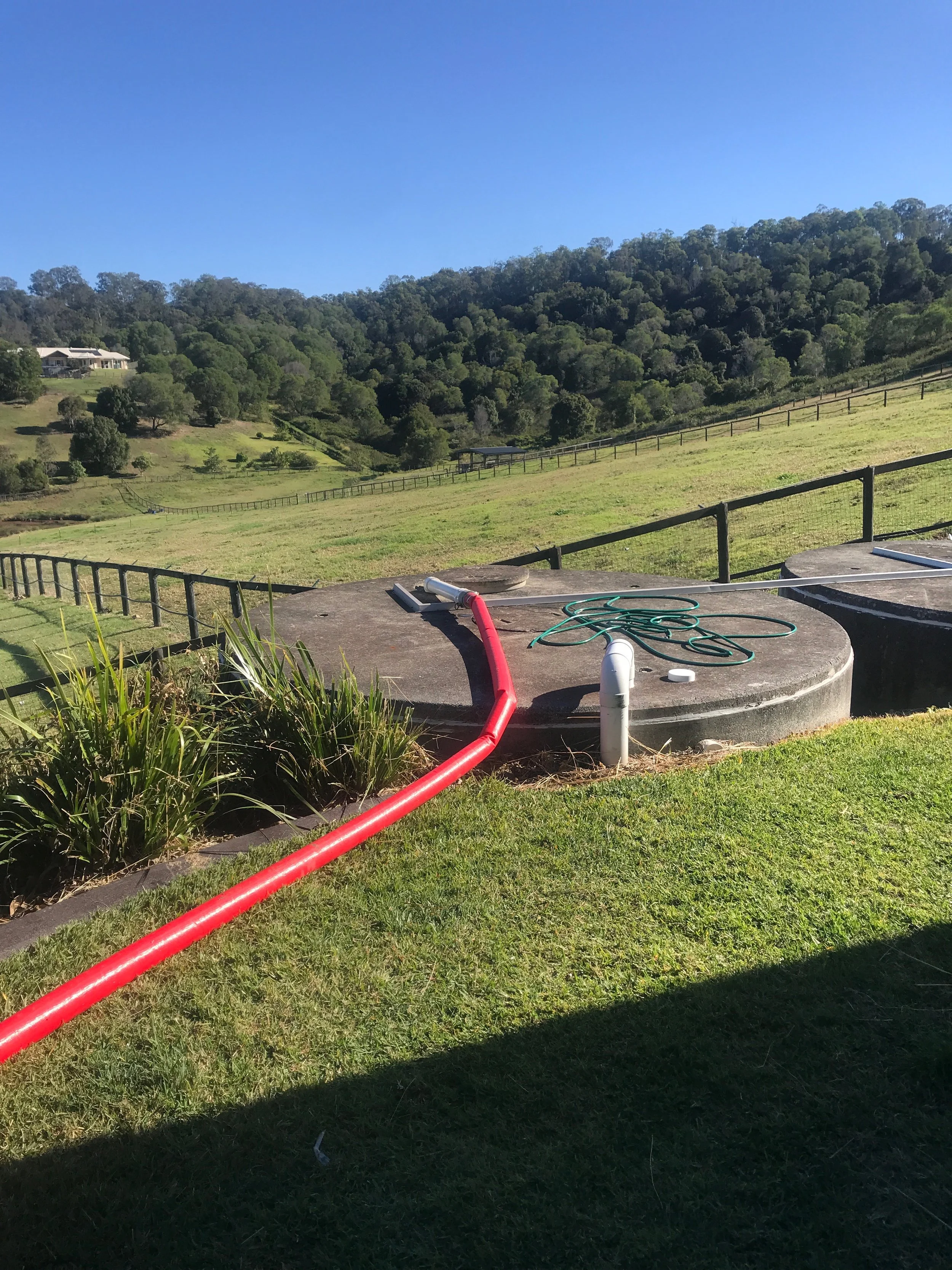 View of a grassy hillside with a fenced yard, some trees, and a house in the distance under a clear blue sky. In the foreground is a concrete water tank with a red hose and a green garden hose on top.