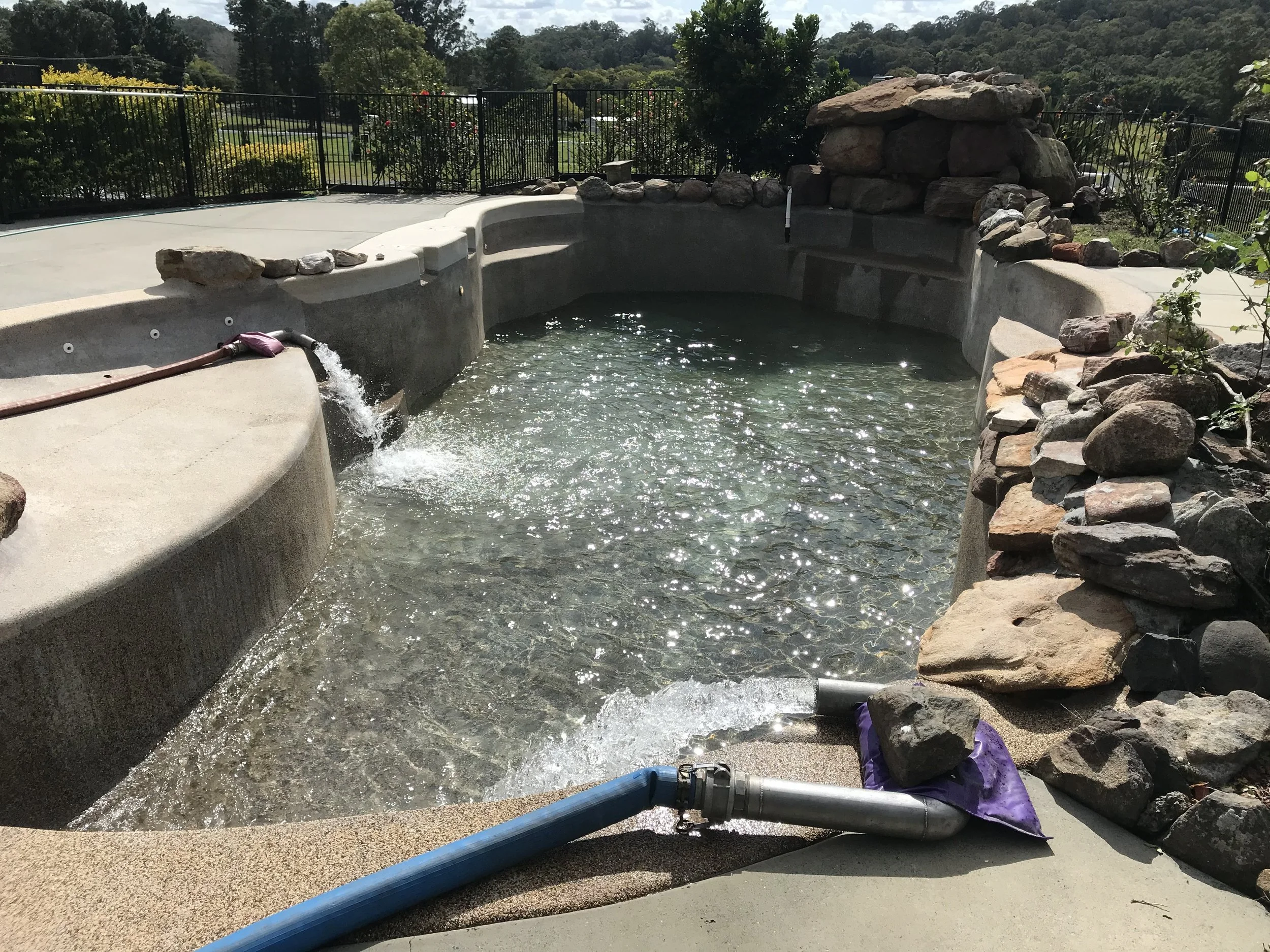Empty hot tub or small pool with rocks and a water spout, during construction or maintenance, on a sunny day with trees and a fence in the background.