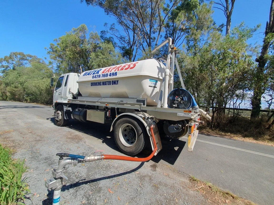 Water tanker truck labeled "Water Free" with a hose connected, parked by a rural road surrounded by trees and blue sky.