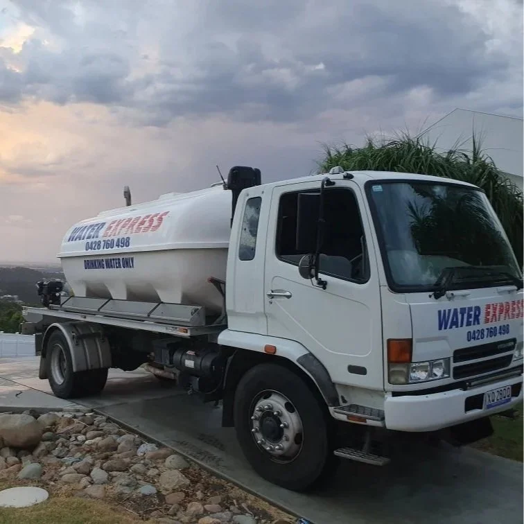 A water delivery truck parked on a driveway with rocks in the foreground, cloudy sky in the background, and a bush to the right.