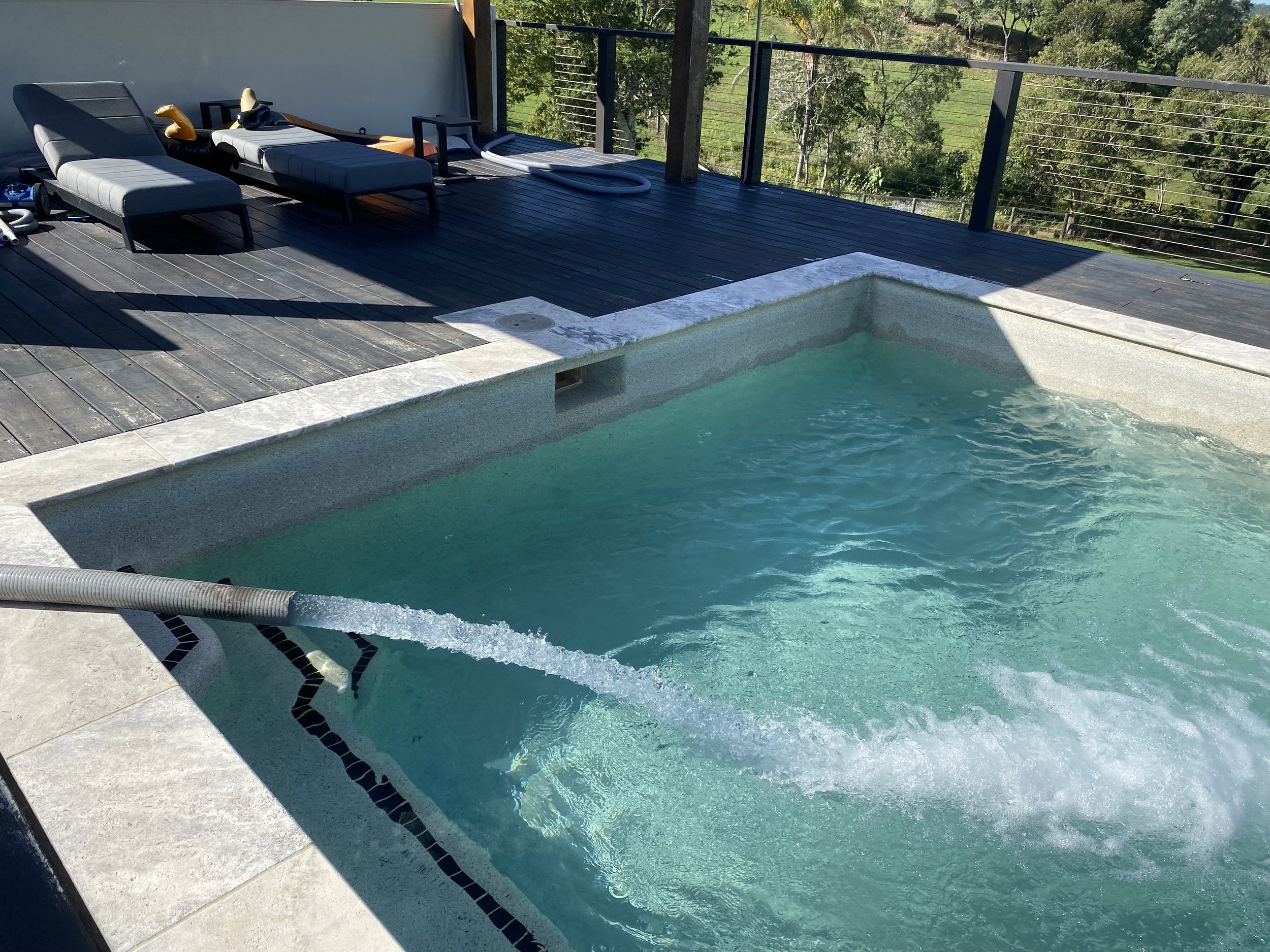A hot tub on a wooden deck with lounge chairs and outdoor furniture, overlooking a green landscape with trees and grass.
