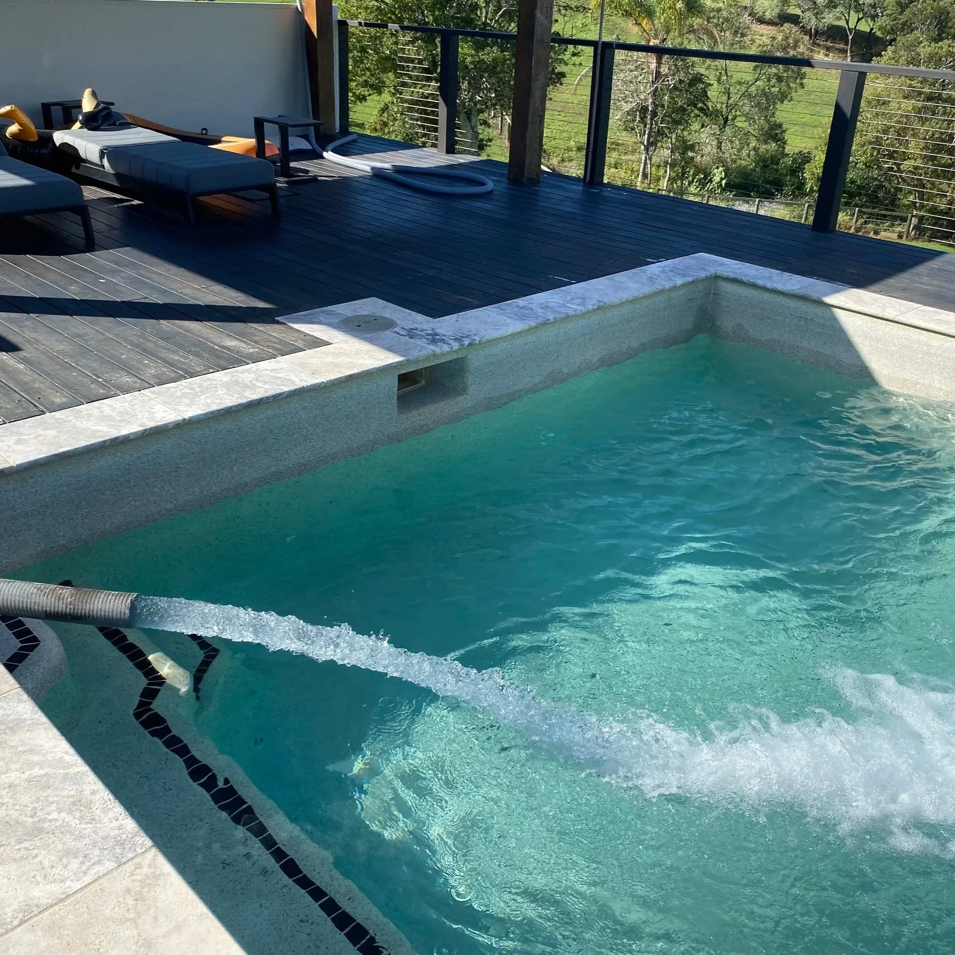 A backyard swimming pool with water jets on a wooden deck, surrounded by trees and greenery.
