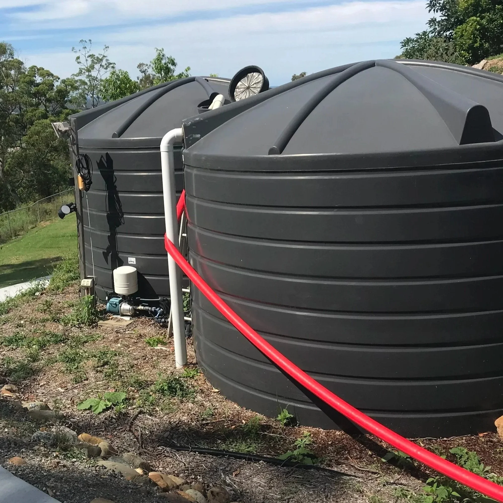 Two large black rainwater tanks on a grassy hillside, with a white pipe and red hose attached.
