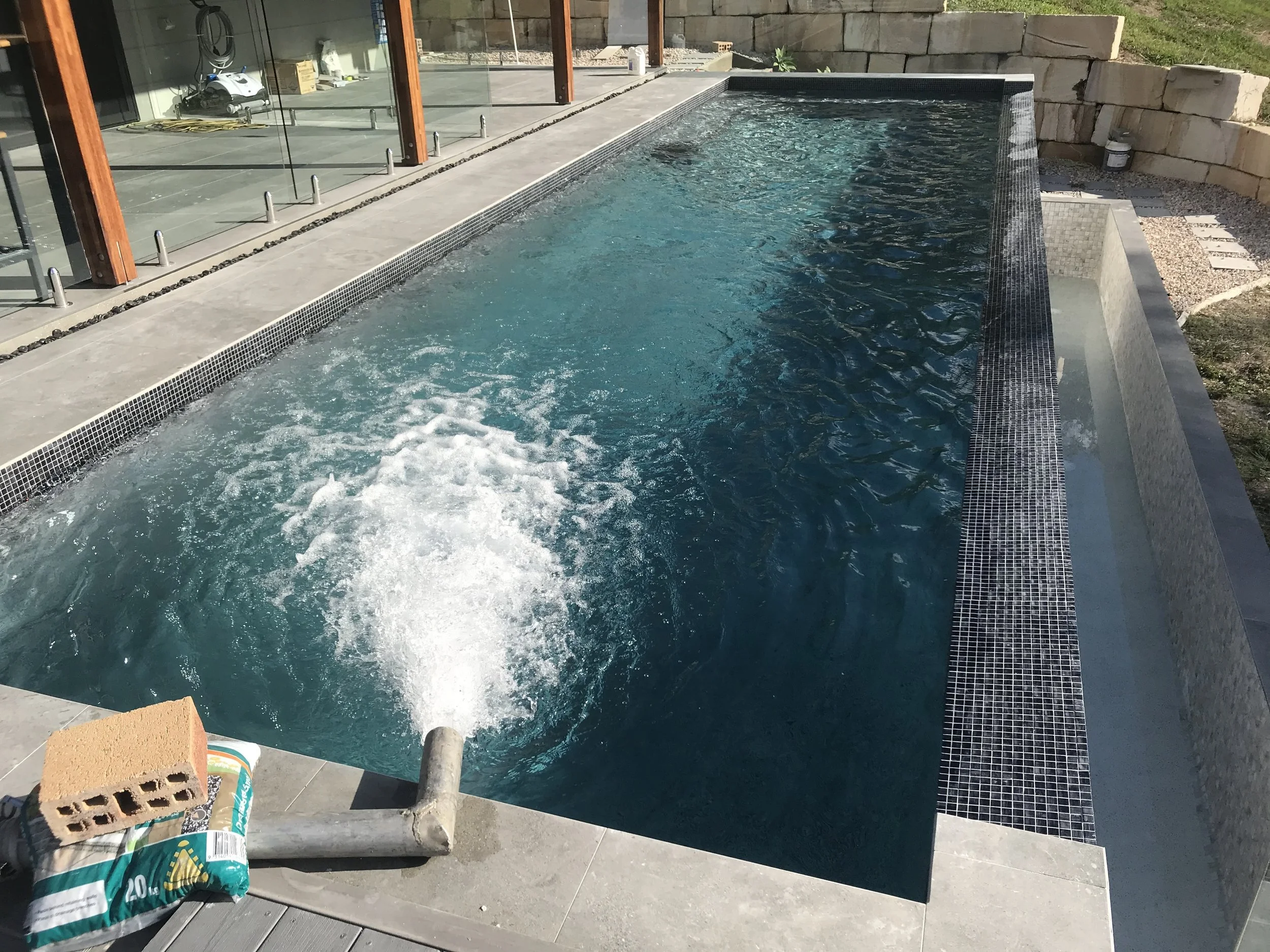 An outdoor rectangular swimming pool with dark mosaic tile edges, partially filled with water, adjacent to a modern house with large glass doors and wooden framing, with a retaining wall of stone blocks and some greenery nearby.