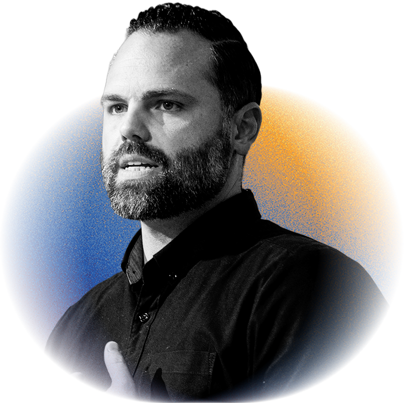 A man with a beard and dark hair, wearing a black shirt, speaking or gesturing, with a blue and yellow circular background.