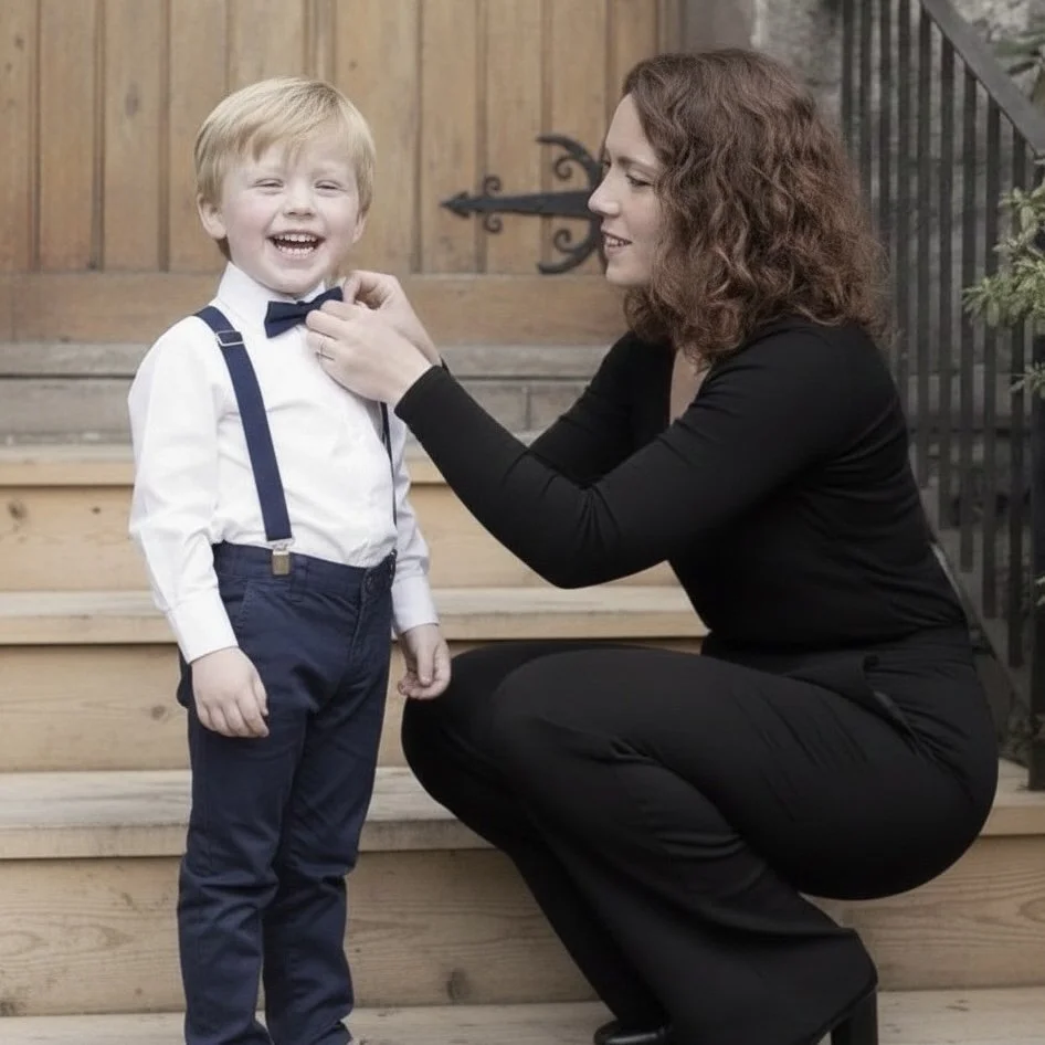 A young boy in a white shirt with suspenders and a bow tie, smiling and standing on steps, while a woman in black, squatting, adjusts his bow tie.