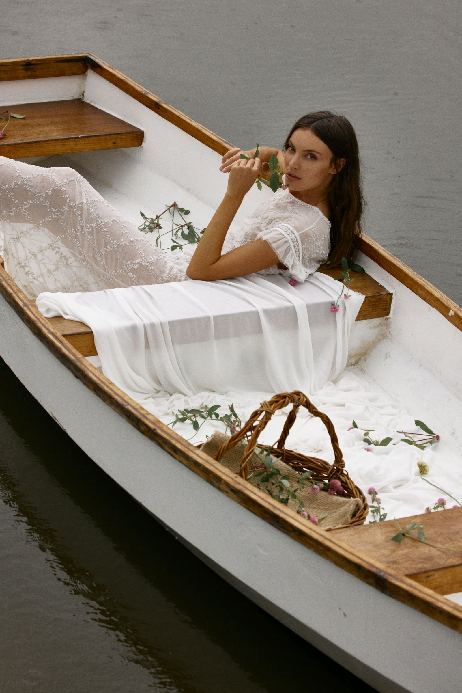 A woman with dark brown hair and light skin lying in a small wooden rowboat on water, wearing a white embroidered dress, holding a leafy branch near her face, with pink flowers scattered in the boat and a wicker basket at the bow.