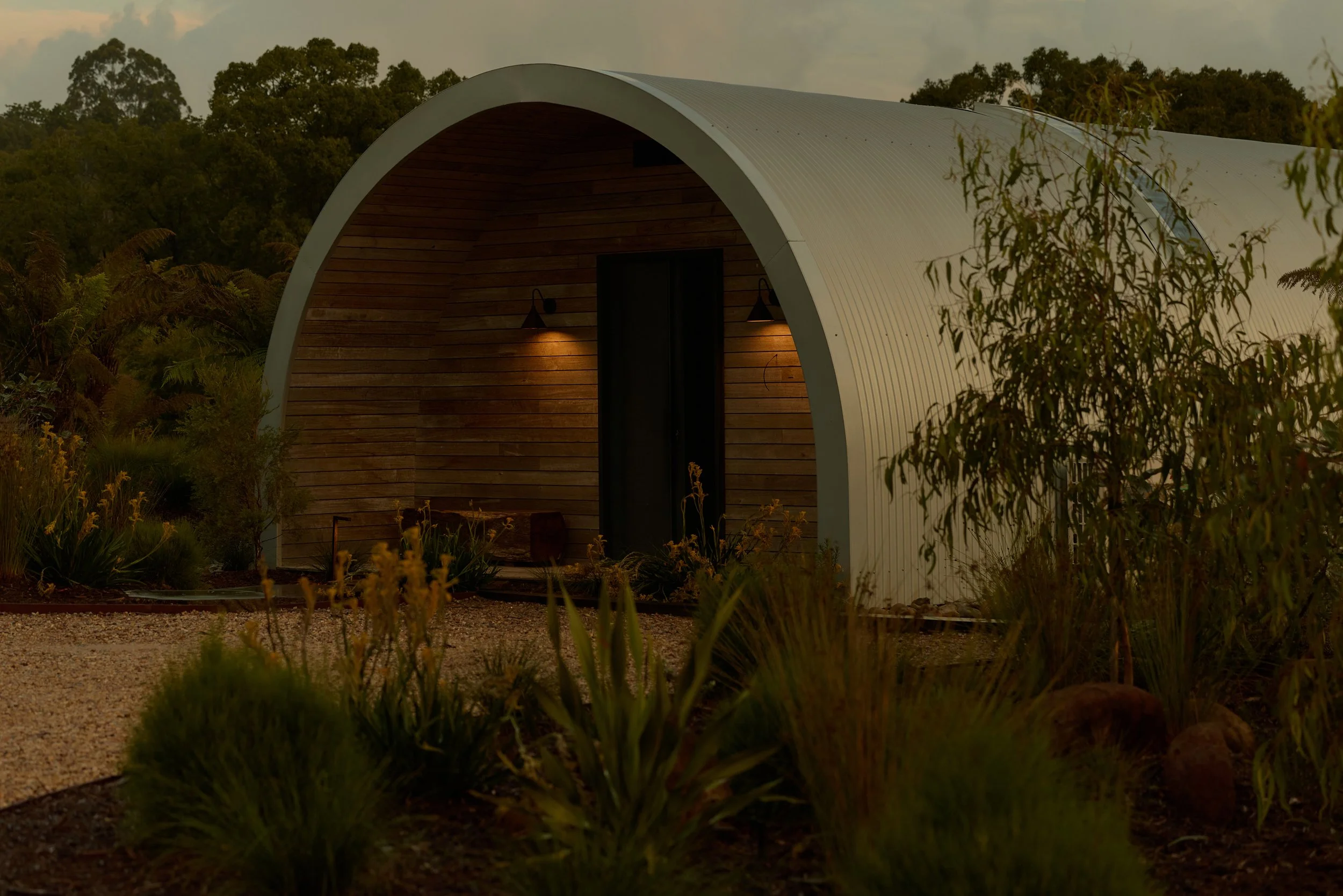 A modern, semi-circular house with a wooden front wall and white curved exterior, surrounded by lush greenery and plants, with soft lighting illuminating the entrance at dusk or evening.