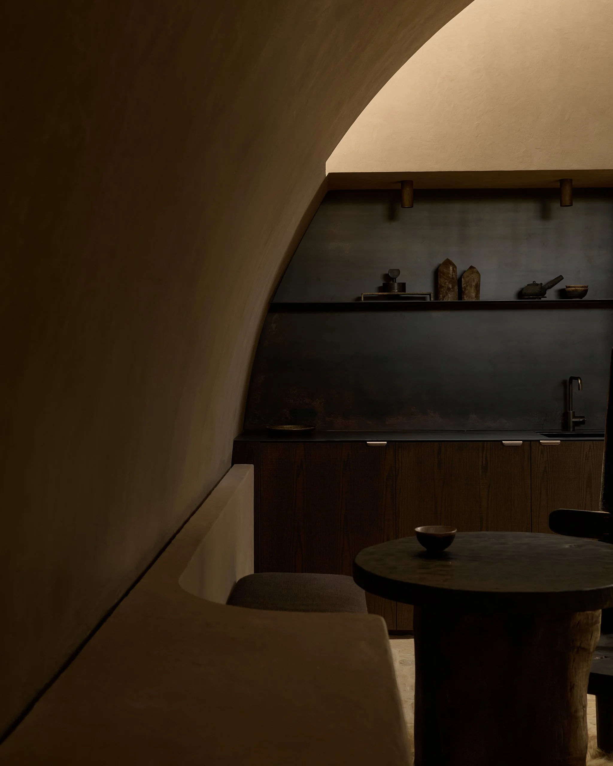 Interior view of a modern minimalist kitchen with a rounded wall, dark wood cabinets, a small table with a bowl, and some decorative items on the counter, with warm lighting.