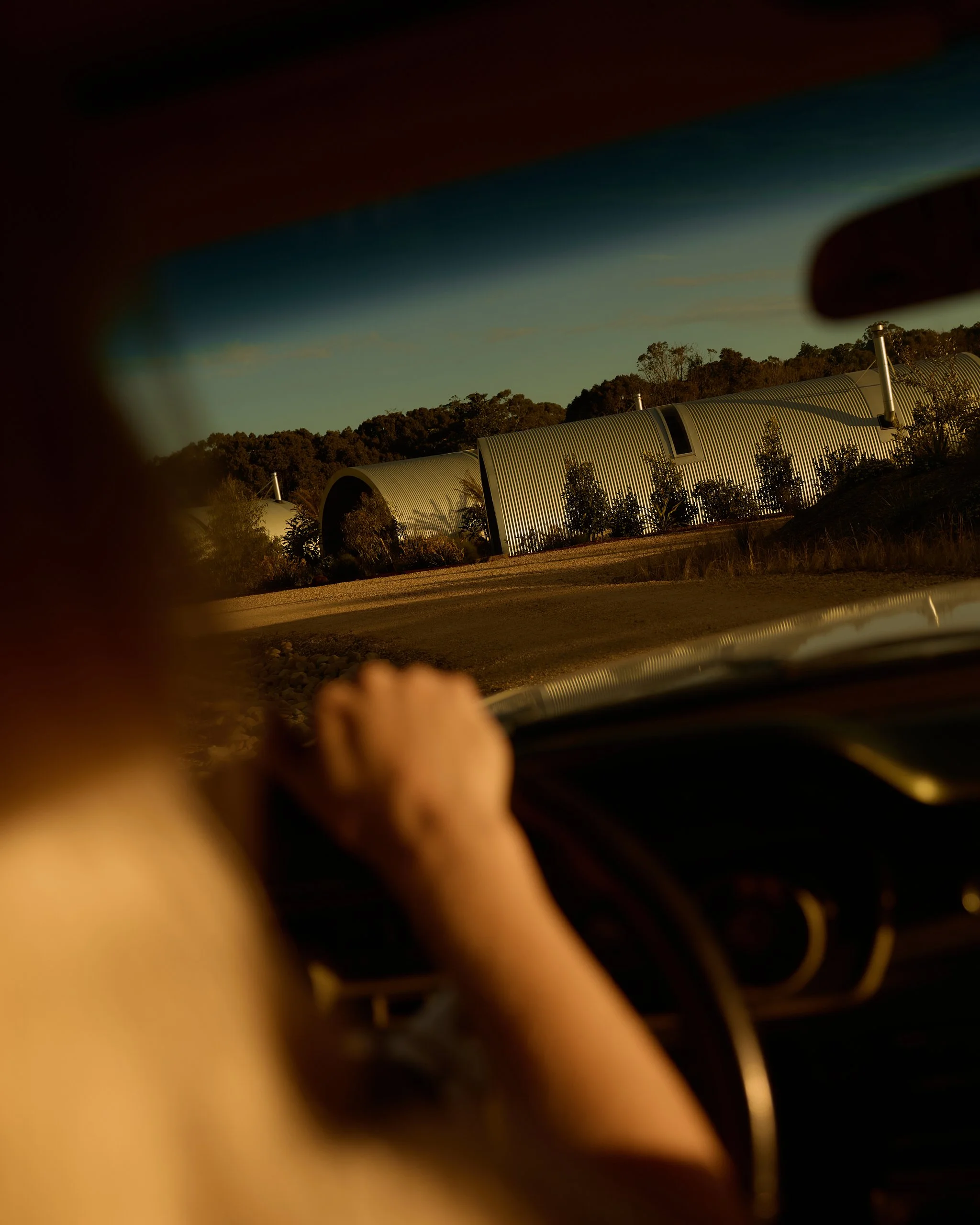 Photo taken from inside a car showing the dashboard, steering wheel, and the view through the windshield of a hilly landscape with metal-roofed buildings and trees under a clear sky.