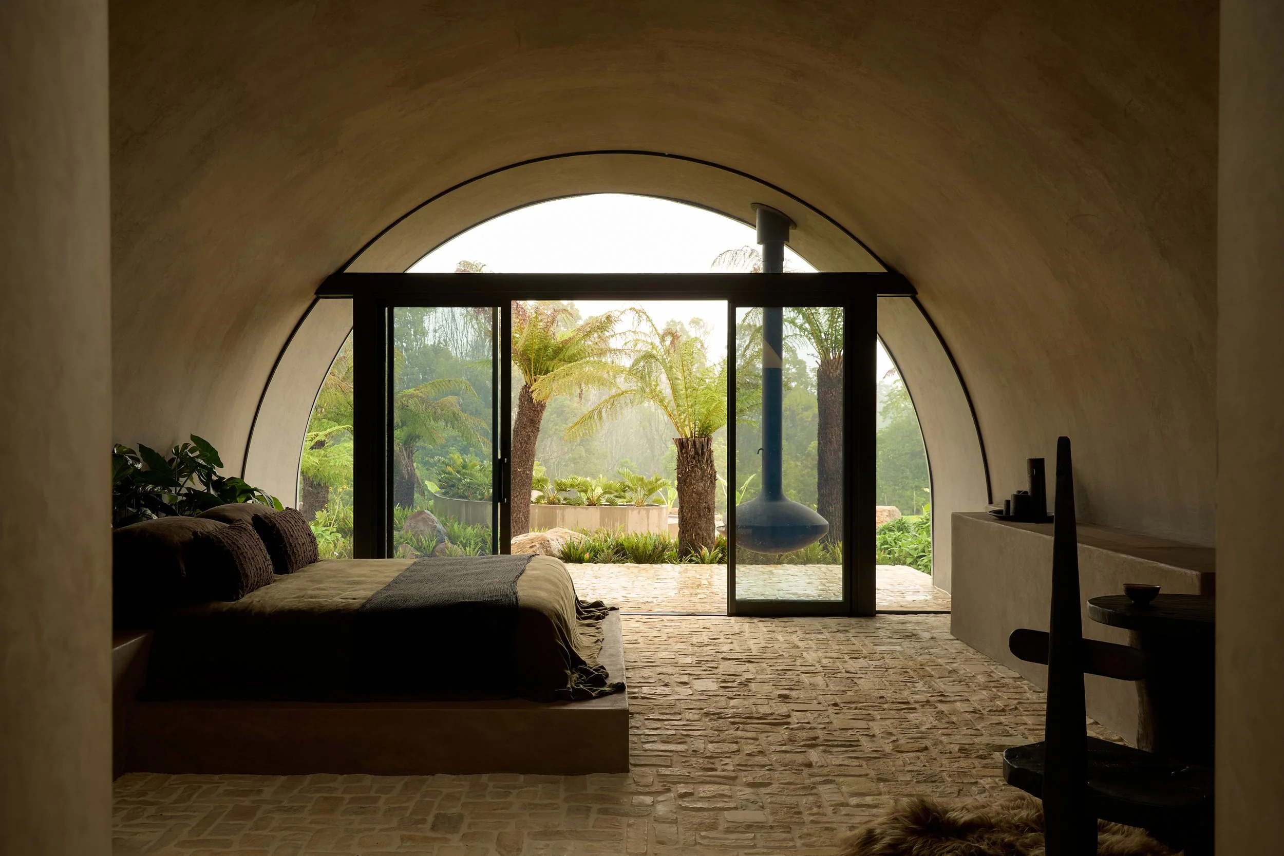 Interior view of a modern, minimalist bedroom with a large arched window opening to a lush garden with trees. The room has a low bed with dark bedding on the left, plants, and a textured stone floor.