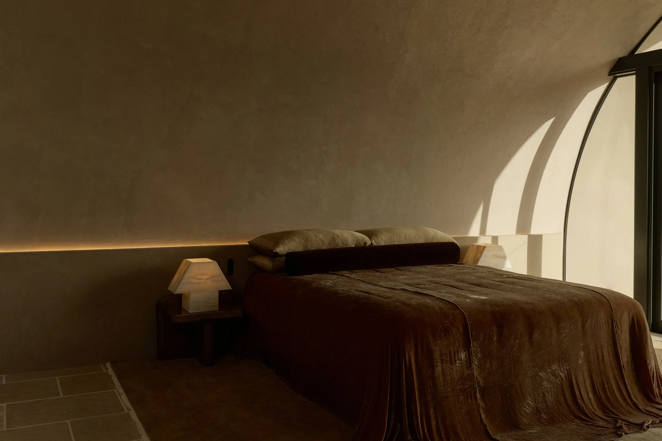 A bedroom with a bed covered in a brown blanket, two pillows, and two bedside lamps with wooden bases. A curved wall with a window and sunlight casting shadows on the wall.