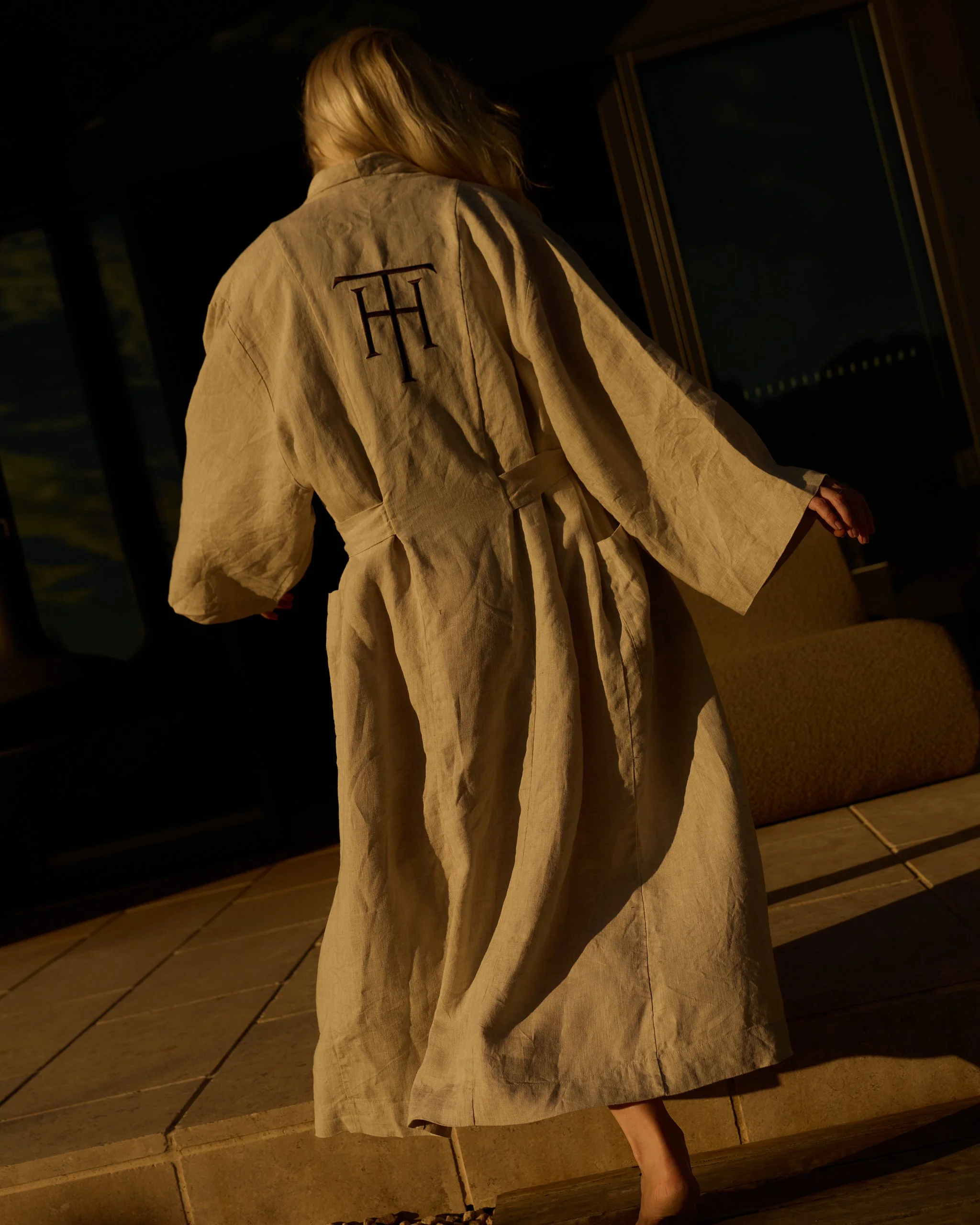Person in a beige robe with a monogrammed letter on the back, standing on a wooden deck at night, facing away from the camera.