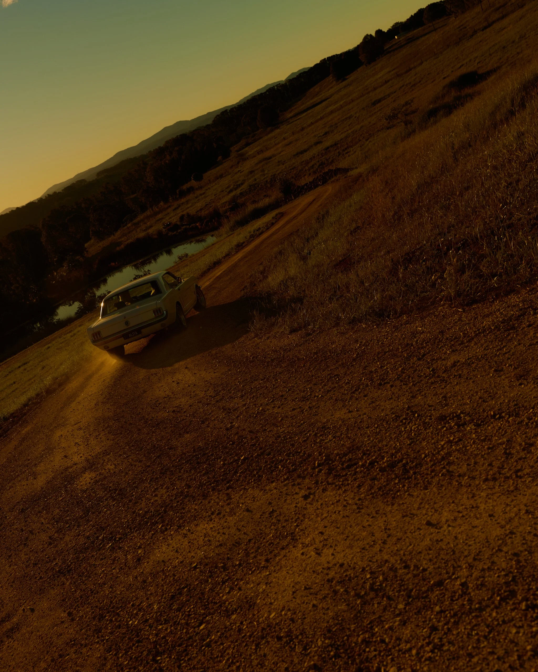 A vintage white car parked beside a small hill with trees, a river, and distant mountains under a sunset sky.