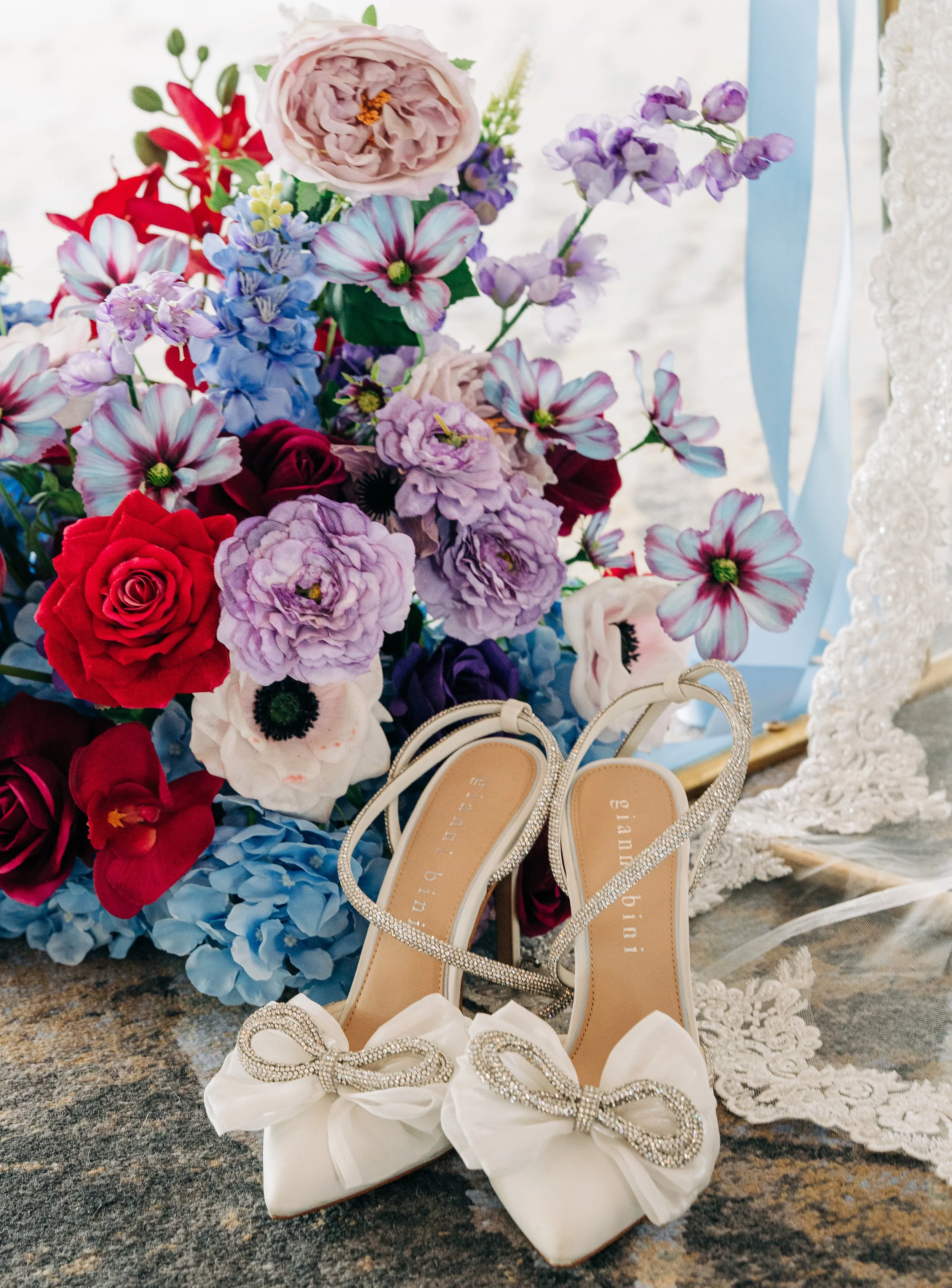 A bouquet of various colorful flowers, including roses, anemones, and delphiniums, next to a pair of white bridal shoes adorned with rhinestone bows, with lace fabric partially visible on the right. Wedding and Events needing a content creator.