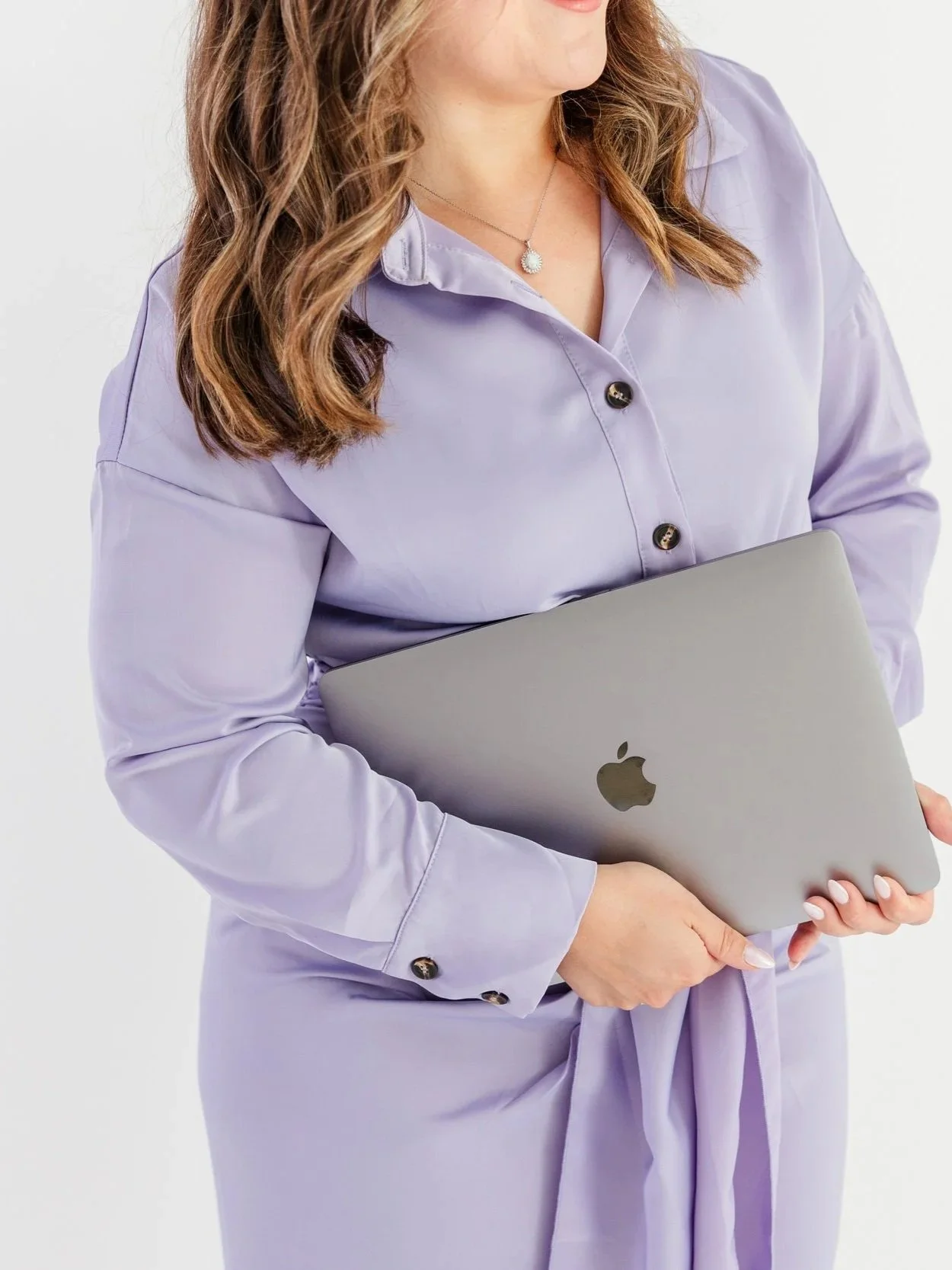 A woman with wavy brown hair wearing a light purple button-up dress, holding a closed silver MacBook laptop.