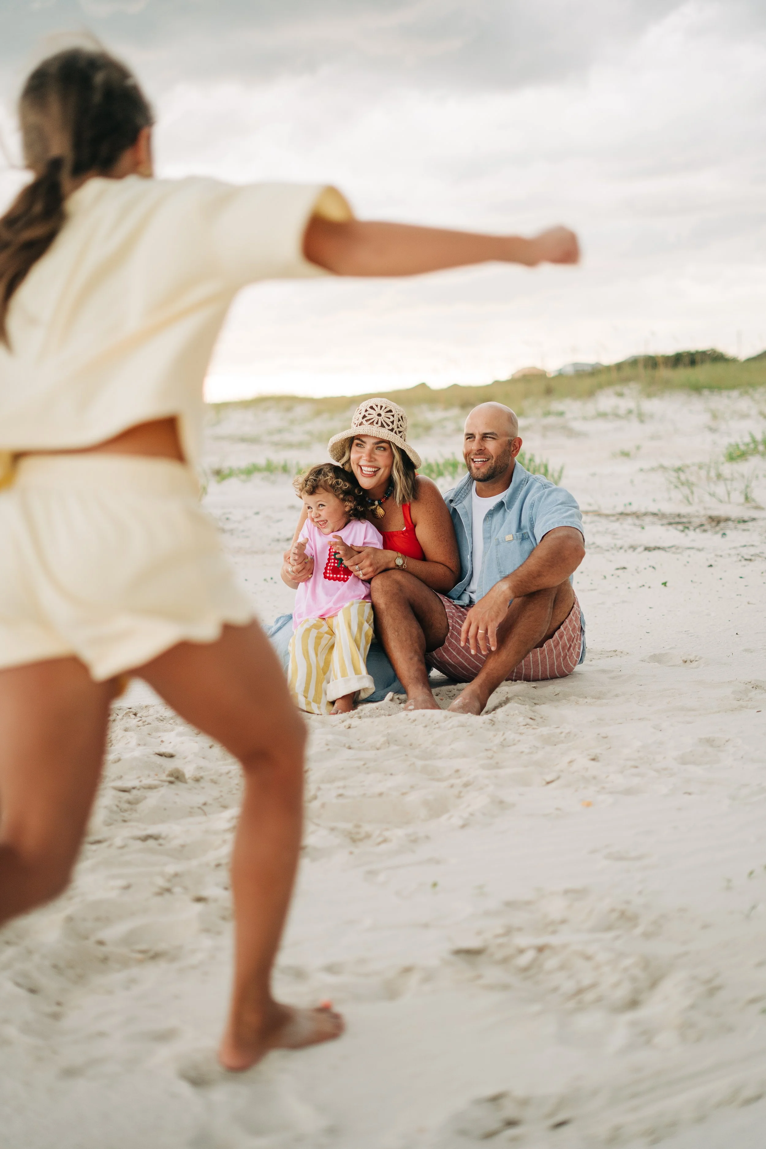 A family of three sitting on the beach, smiling and enjoying time together as a young girl and adult woman are playing in front. Family Photography in South Alabama.