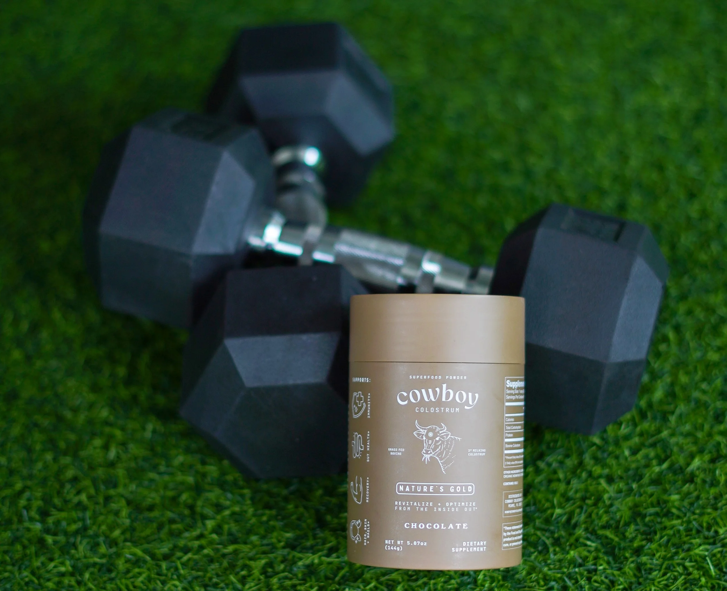 A container of Cowboy Colostrum supplement in front of a pair of black dumbbells on green artificial grass.