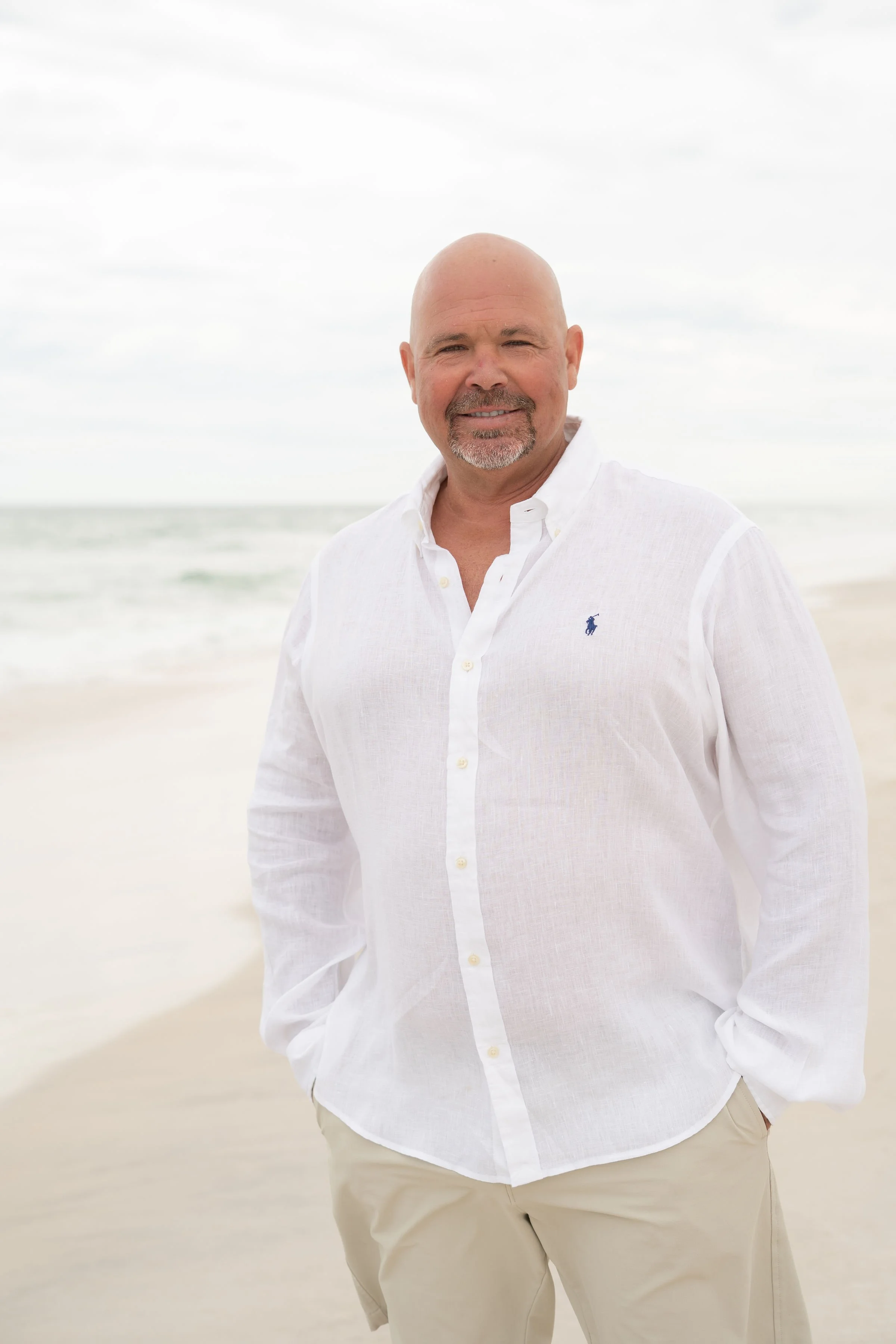 A smiling, bald man with a goatee, wearing a white button-up shirt with a small logo, standing on a sandy beach with the ocean and sky in the background.