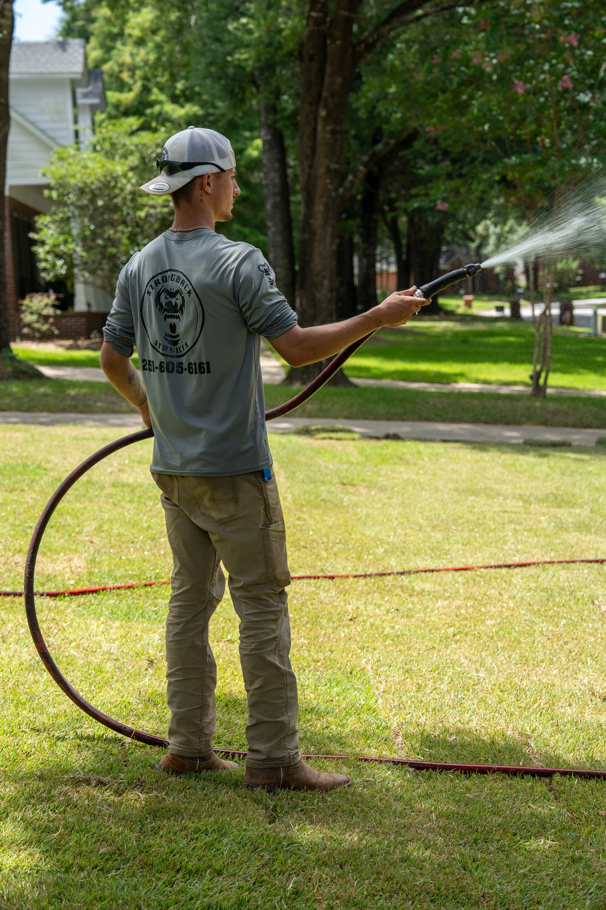 A man wearing a gray long sleeve shirt, khaki pants, and a baseball cap is watering a lawn in a residential neighborhood. The man’s shirt has a logo and phone number on the back. Job Site Marketing photos in Fairhope, Spanish Fort, and Daphne.