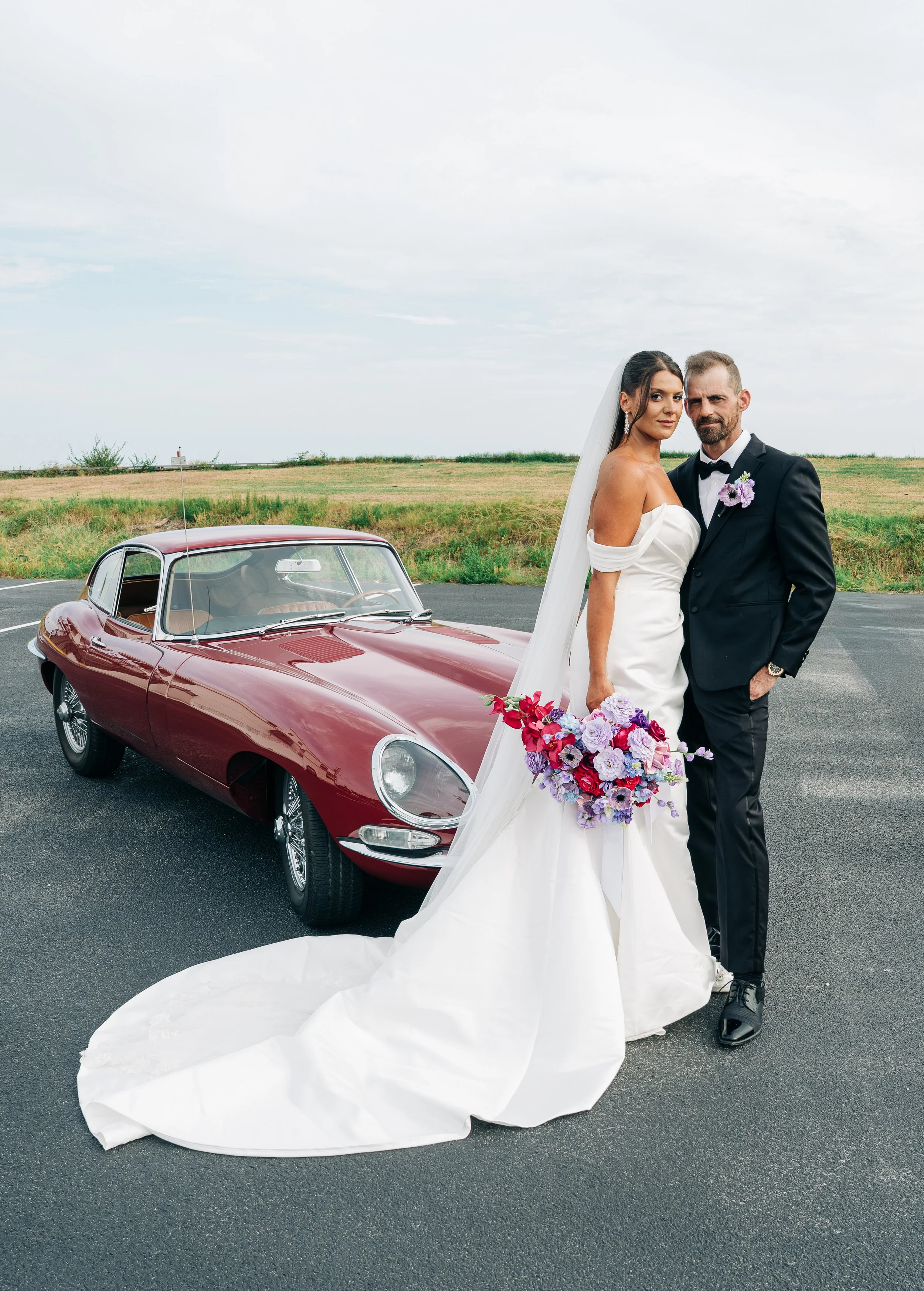 A bride and groom standing next to a vintage red sports car in an open field. The bride is wearing a white off-shoulder wedding gown and holding a large bouquet of colorful flowers. The groom is dressed in a black tuxedo with a bow tie and boutonniere. They are looking at the camera with serious expressions. The sky is partly cloudy.