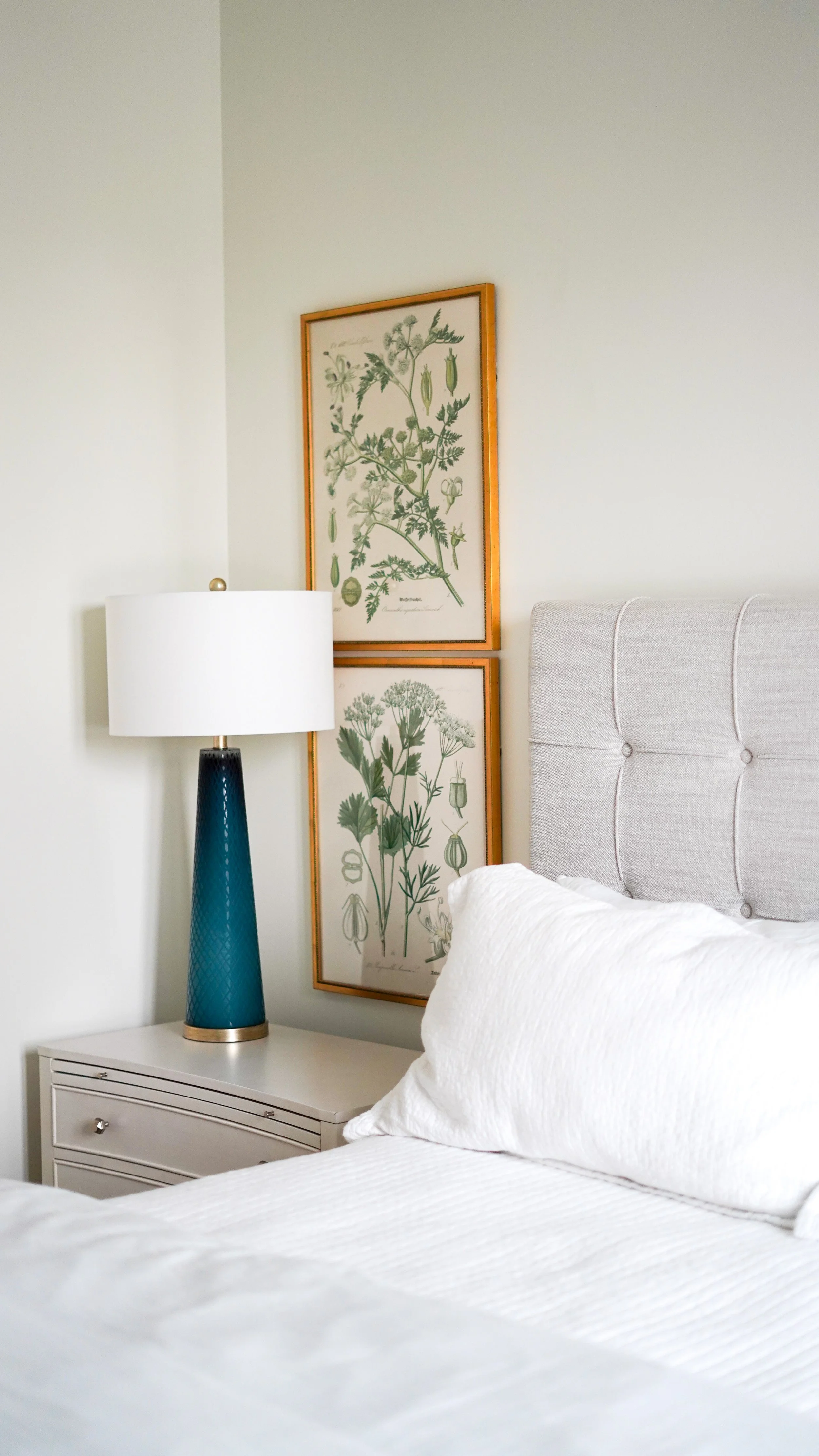 A bedroom corner with a white bed, a gray headboard, a nightstand, a blue table lamp, and framed botanical prints on the wall.