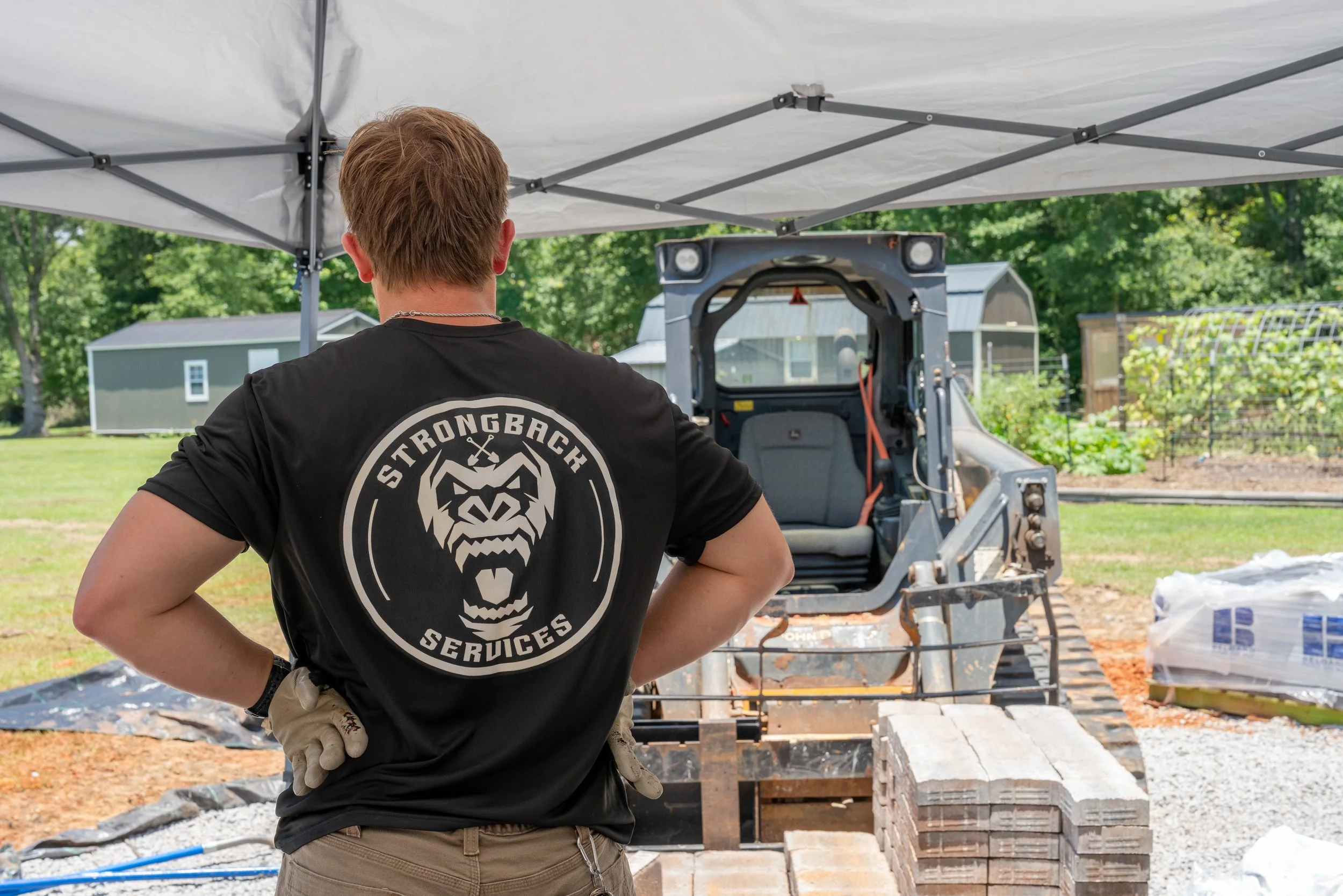 A person with short brown hair wearing a black t-shirt with a logo on the back, standing outdoors under a canopy, observing construction work involving bricks and machinery in a backyard. Content Creation for business owners on the Alabama Gulf Coast