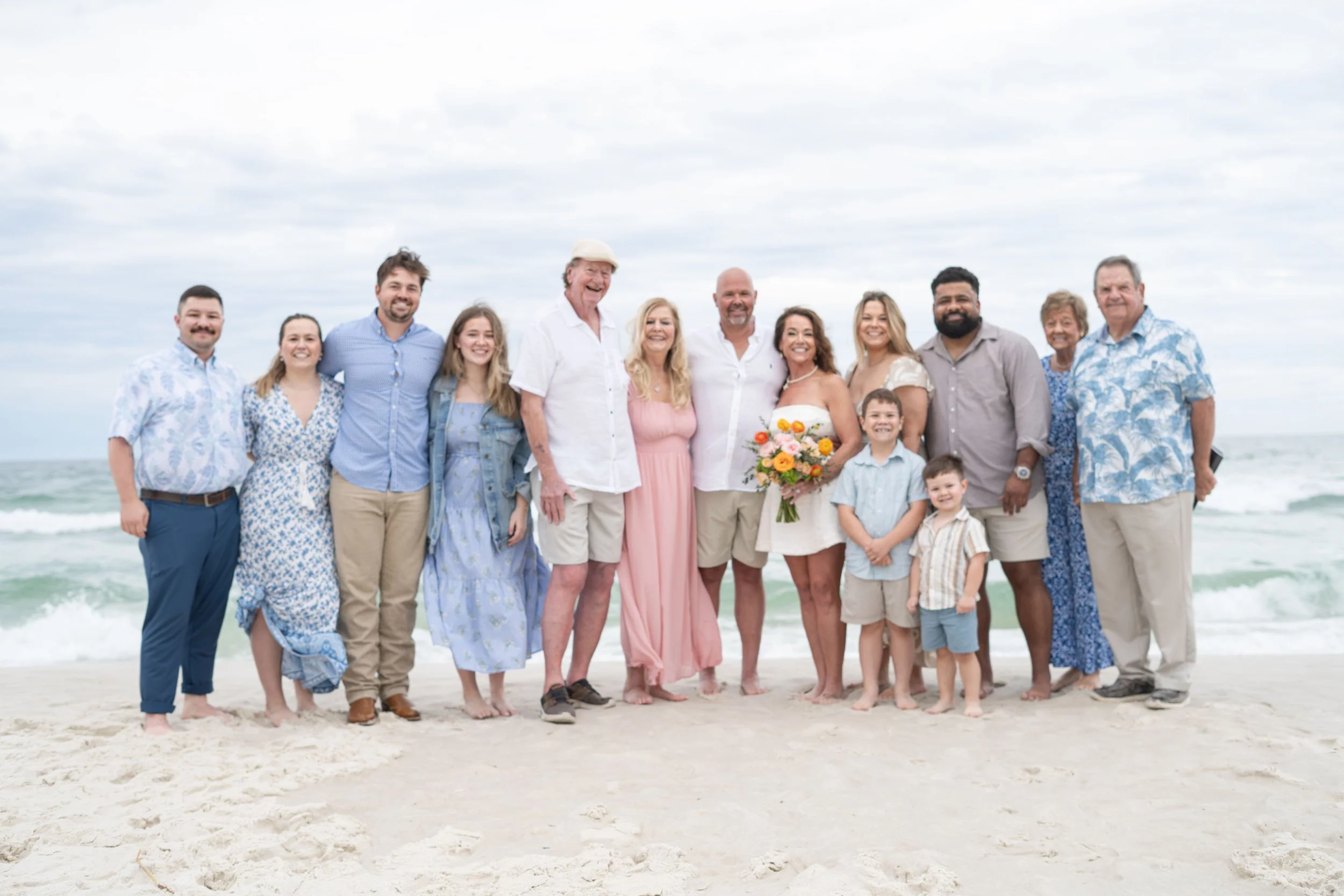 A large family standing on the beach with the ocean in the background. They are dressed in casual, summer clothing and smiling, with a woman holding a bouquet of flowers.