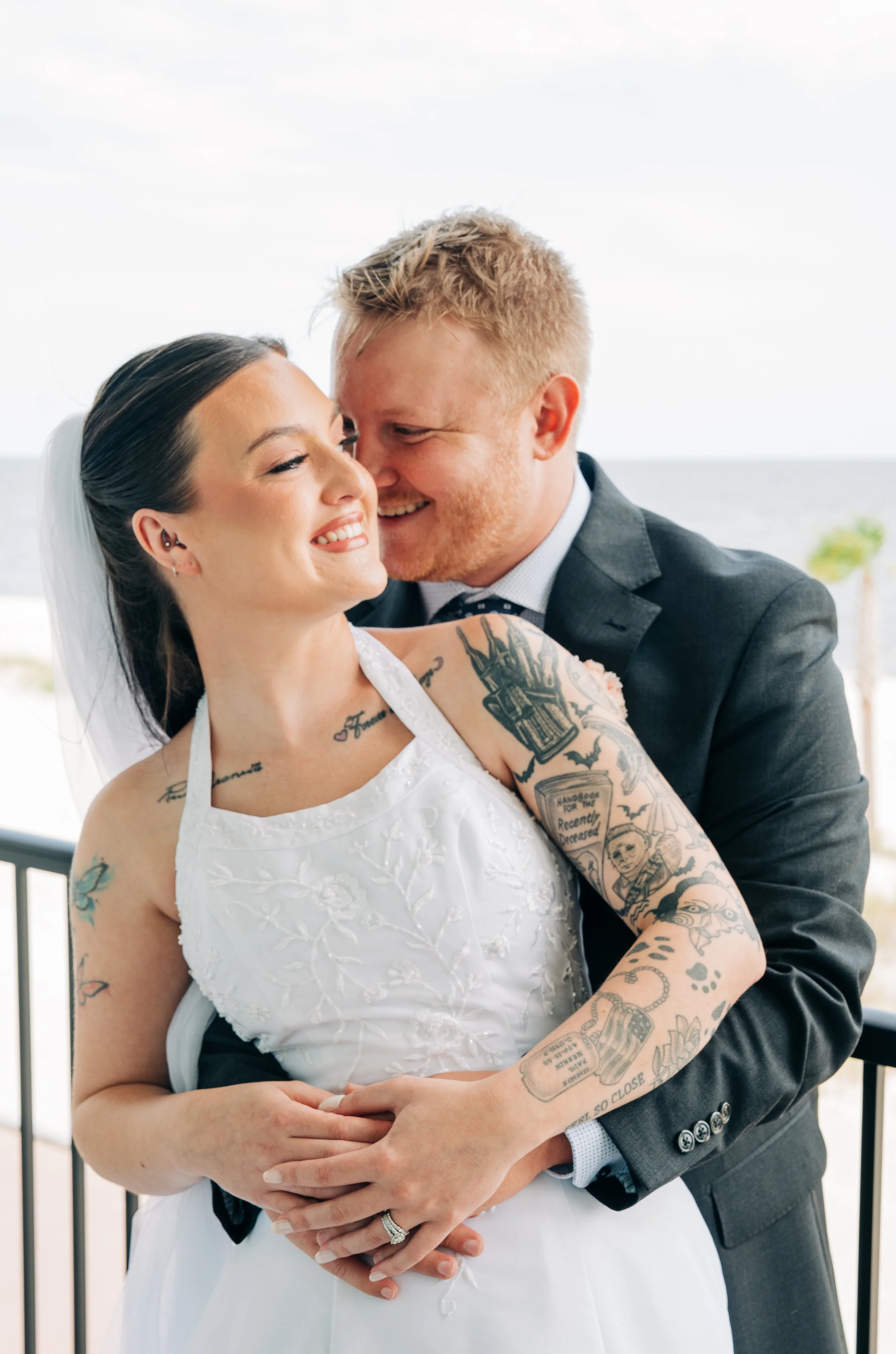 A bride and groom are embracing on a balcony, smiling at each other with the ocean in the background. Wedding photos.