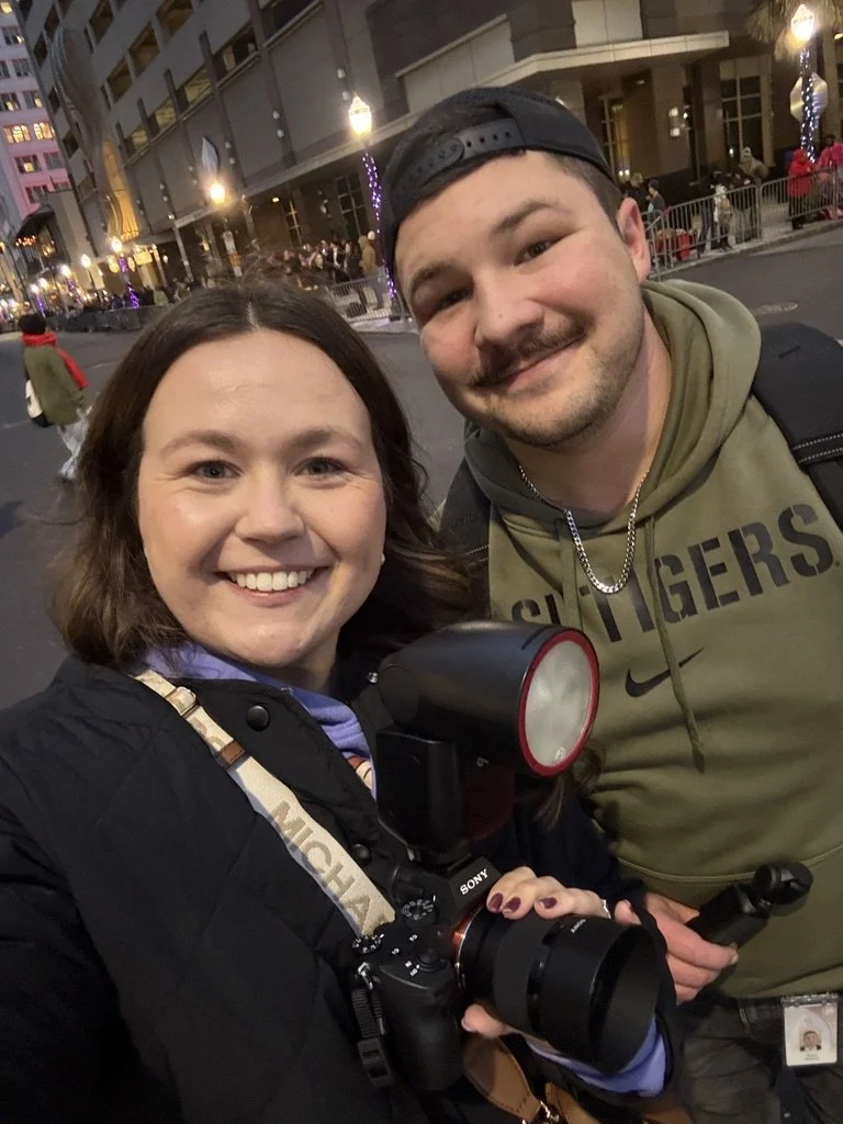 A woman and a man smiling for a selfie outside in an urban setting at night. The woman is holding a camera with a large flash attachment, and the man is wearing a hoodie, a backward cap, and a chain necklace. In the background, there are buildings, streetlights, and people walking along the sidewalk.